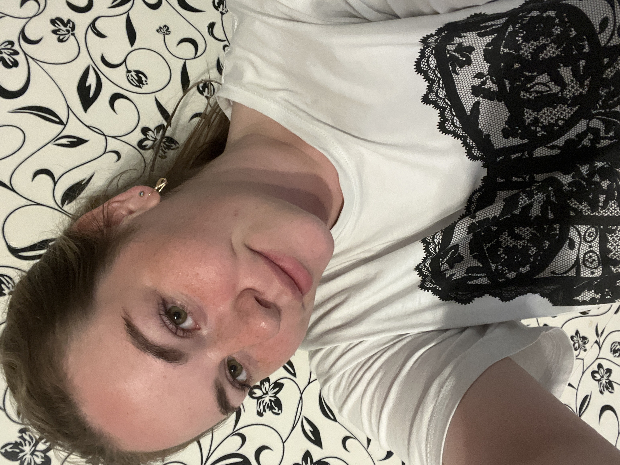 The image shows a woman with light skin, freckles, and green eyes. She is lying on a surface with a black and white floral pattern, possibly a bed or a couch. She is wearing a white t-shirt with black lace details. The location is potentially Chaikovsky, Russia, but this cannot be confirmed solely based on the visual information in the image.