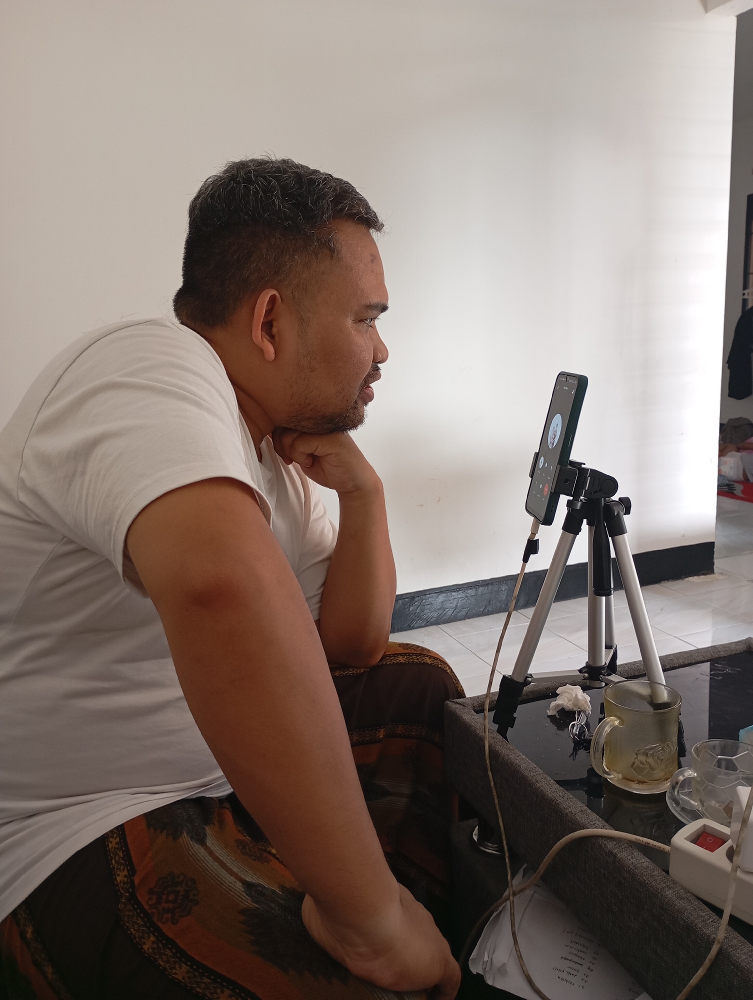 The image depicts a man, likely of Indonesian ethnicity, seated and facing a smartphone mounted on a tripod. The man, with short, graying hair and a beard, is wearing a white t-shirt and patterned pants, possibly a sarong. His right hand is supporting his chin, and he appears to be engaged in a video call. The smartphone screen displays a video call interface with a profile picture and call controls.
The setting appears to be an indoor space, likely a living room, with a white wall in the background. In the foreground, a dark-colored coffee table holds a glass mug with liquid, another empty glass, a small pile of crumpled paper, and a power strip with a switch. Cables are visible, suggesting the smartphone may be charging. The floor is tiled.
The overall impression is that of a person engaged in a virtual communication session in a domestic environment. The presence of personal items and the casual attire suggest a relaxed, personal setting. The image is likely taken during daylight hours, given the lighting conditions.
While the image provides no explicit identifiers of Mataram, Indonesia, the subject's likely ethnicity, clothing style, and the overall ambiance are consistent with a domestic scene in Indonesia. The presence of what appears to be a sarong further strengthens this assumption.