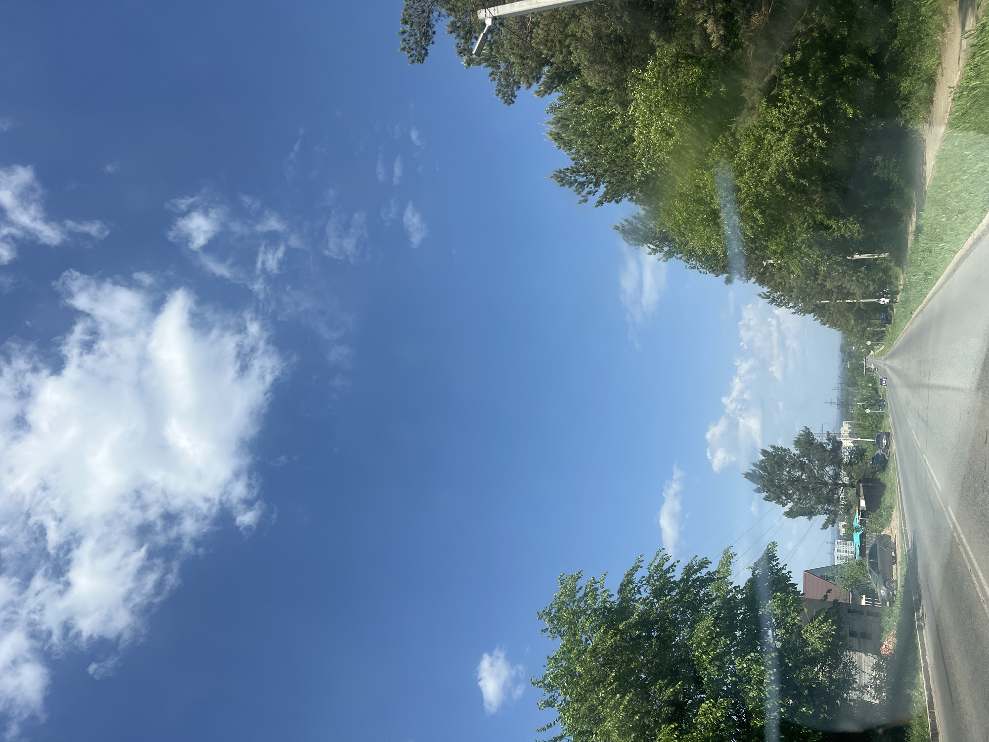 The image shows a view from a vehicle traveling on a road in what appears to be a suburban area. The sky is predominantly blue with scattered white clouds. Trees line both sides of the road, forming a canopy. In the distance, buildings and power lines are visible. Based on the context provided, this location is likely in or near Chaikovsky, Russia. The road surface is asphalt, and there is a shoulder or grassy area adjacent to the road.