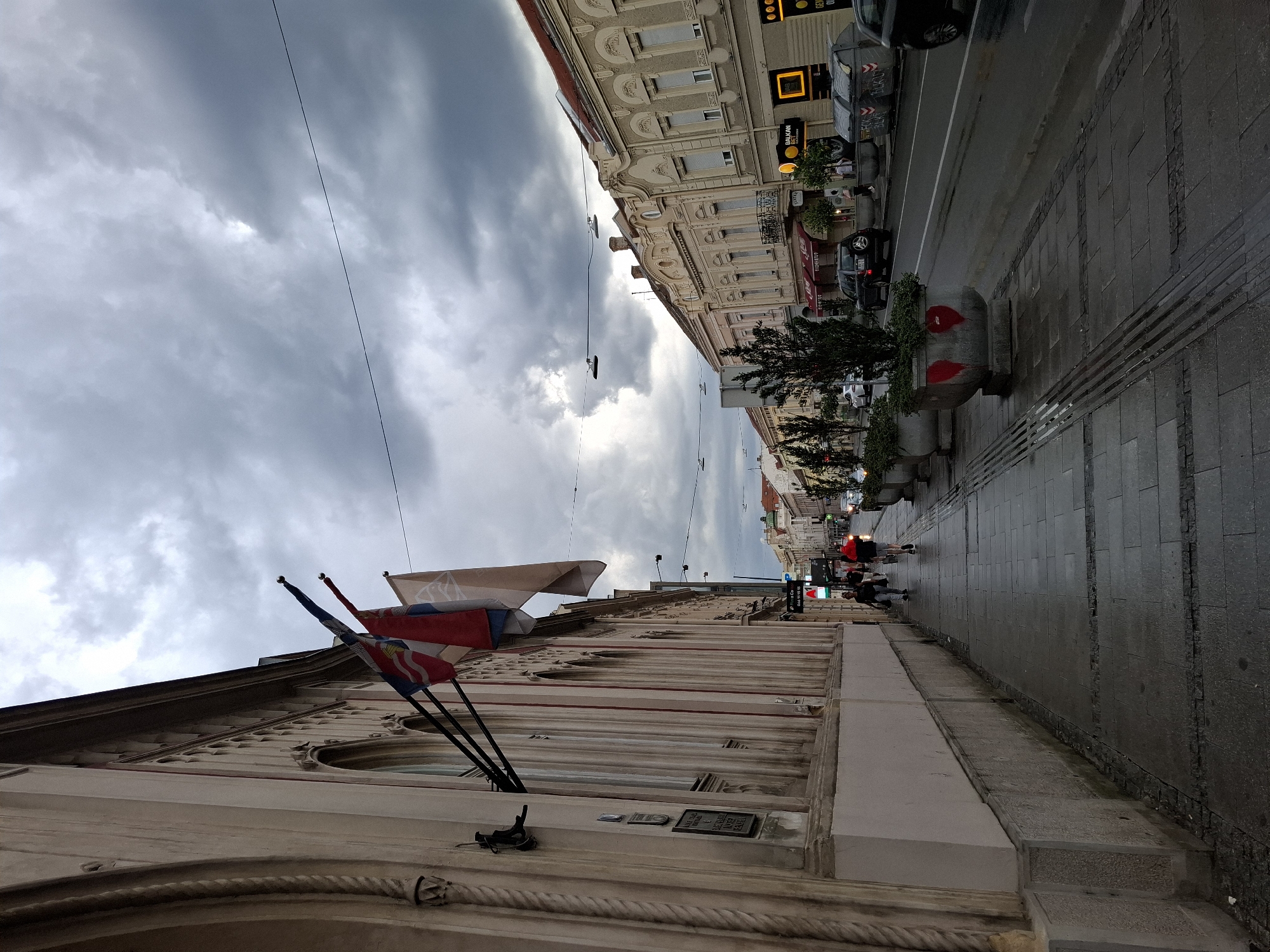 The image depicts a street scene in Zemun, Serbia, under a cloudy sky. On the left, the facade of a building with arched windows and ornate trim is visible, adorned with several flags. A sidewalk with patterned paving stones runs along the building. Several individuals are walking on the sidewalk. To the right of the sidewalk is a street with a lane marking, wet from rain. Several cars are parked or driving on the street. The street is lined with buildings with similar architectural styles, featuring decorative facades and awnings. Large concrete planters with trees are placed along the sidewalk. A sign with "BALKAN" is visible on one of the buildings. Several large trash bins are positioned near the curb. The overall atmosphere is overcast and damp.