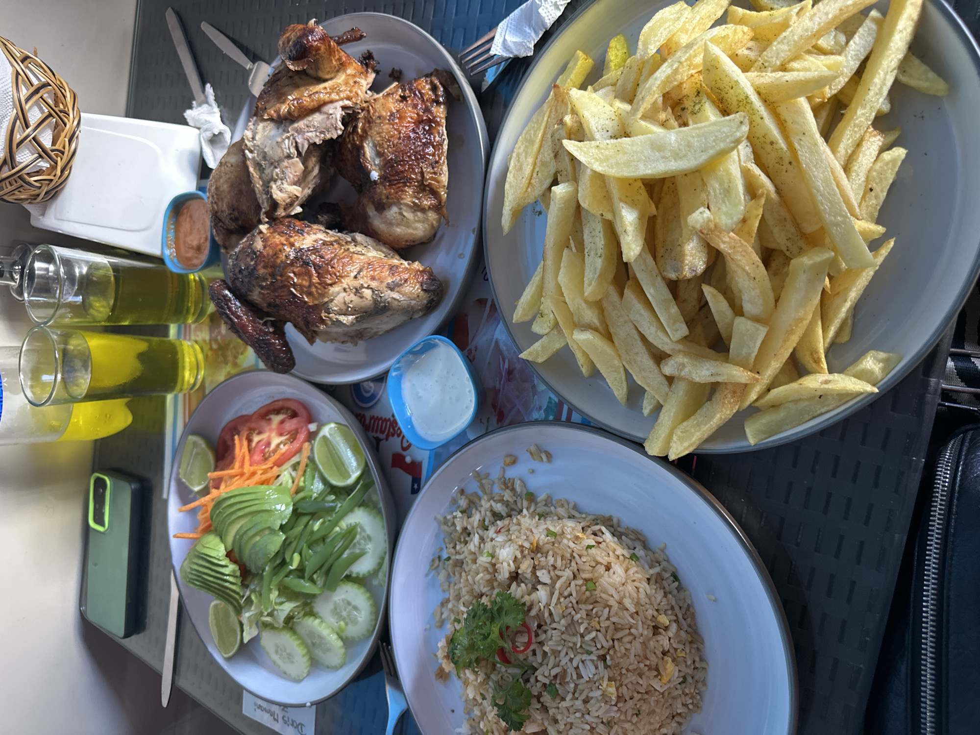 The image depicts a meal scene, likely outdoors in Arica, Chile, given the plates marked with "Restaurante 7 Mares - Arica." Four plates are displayed: one with roasted chicken pieces, one with french fries, one with a salad of avocado, tomatoes, cucumber, shredded carrots, and limes, and one with fried rice. Small condiment containers, possibly with sauces, are also visible. A basket, cutlery, and two bottles of olive oil (or similar) add to the setting. An olive green smartphone lies near the salad plate, while a black leather bag is visible to the right. The meal is presented on a textured gray surface.
