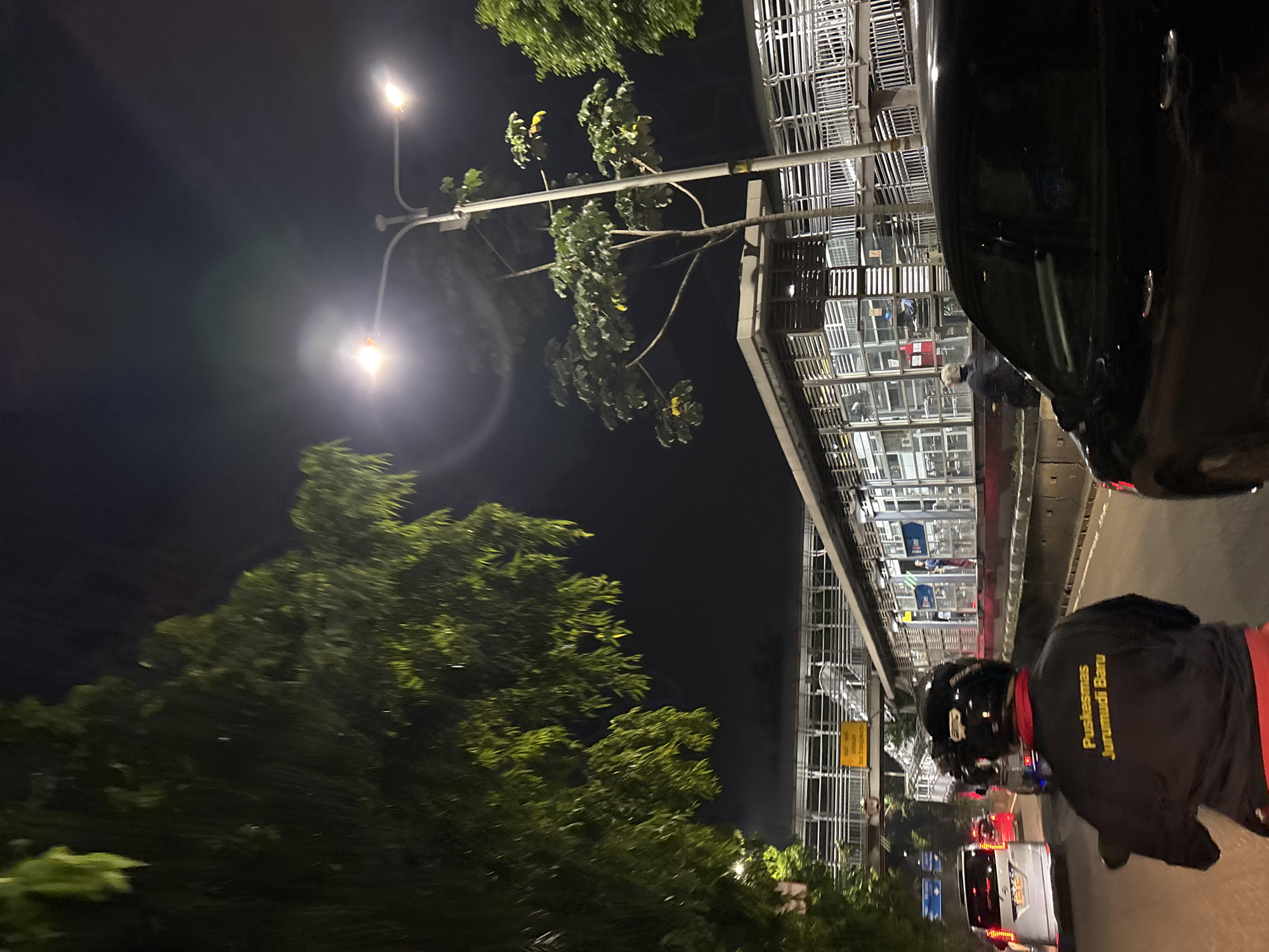 The image depicts a street scene at night in West Jakarta, Indonesia, likely near a public transportation hub. The foreground is dominated by a person riding a motorcycle, viewed from behind. The rider is wearing a black jacket with yellow text that reads "Puskesmas Jurumudi Baru," indicating a health center. The motorcycle has a black helmet on the rider. To the right of the motorcycle, a portion of a black car is visible.
The middle ground of the image features a busy street with several vehicles, including a white car and a red car. Beyond the road, there's a modern building with glass facades and metal railings, possibly a bus stop or a train station. Streetlights illuminate the area. Trees with green leaves are visible on the left side of the image, contrasting with the dark night sky. The overall environment suggests an urban setting with ongoing traffic and illuminated infrastructure.