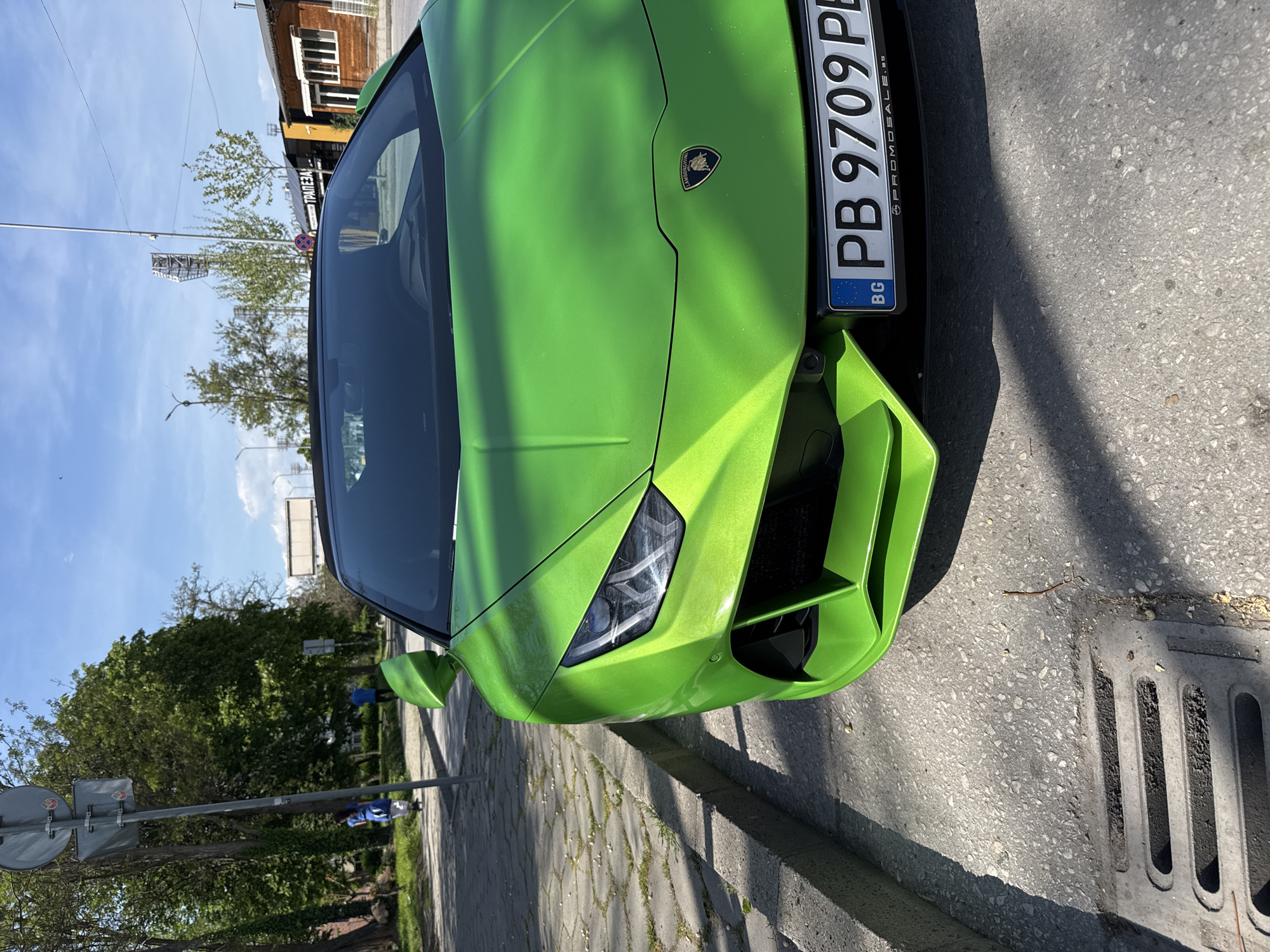 The image, rotated 90 degrees clockwise, prominently features a vibrant, almost neon green Lamborghini Huracan parked on a paved street. The car occupies the majority of the frame, showcasing its sleek front design, including the distinctive headlights, air intakes, and the iconic Lamborghini emblem on the hood.
The front Bulgarian license plate, oriented vertically on the car's right, displays "BG" and the registration "PB 9709 PL". The "PB" code confirms the location as Plovdiv, Bulgaria. The license plate frame also includes the text "PROMOSALE" and "BG".
The scene is set outdoors on a sunny day, indicated by a clear blue sky and strong shadows cast by the car and unseen objects onto the pavement, suggesting bright daylight, possibly mid-morning or afternoon. In the background, a brown wooden building with a sign displaying the Bulgarian word "ТРАПЕЗА" (TRAPEZA, meaning 'table' or 'meal') is visible. Other background elements include various trees with green foliage, utility poles, and street signs. The street itself consists of asphalt and concrete paving stones, with a metal drain grate visible towards the bottom right of the image. No people are visible, and the car appears stationary.