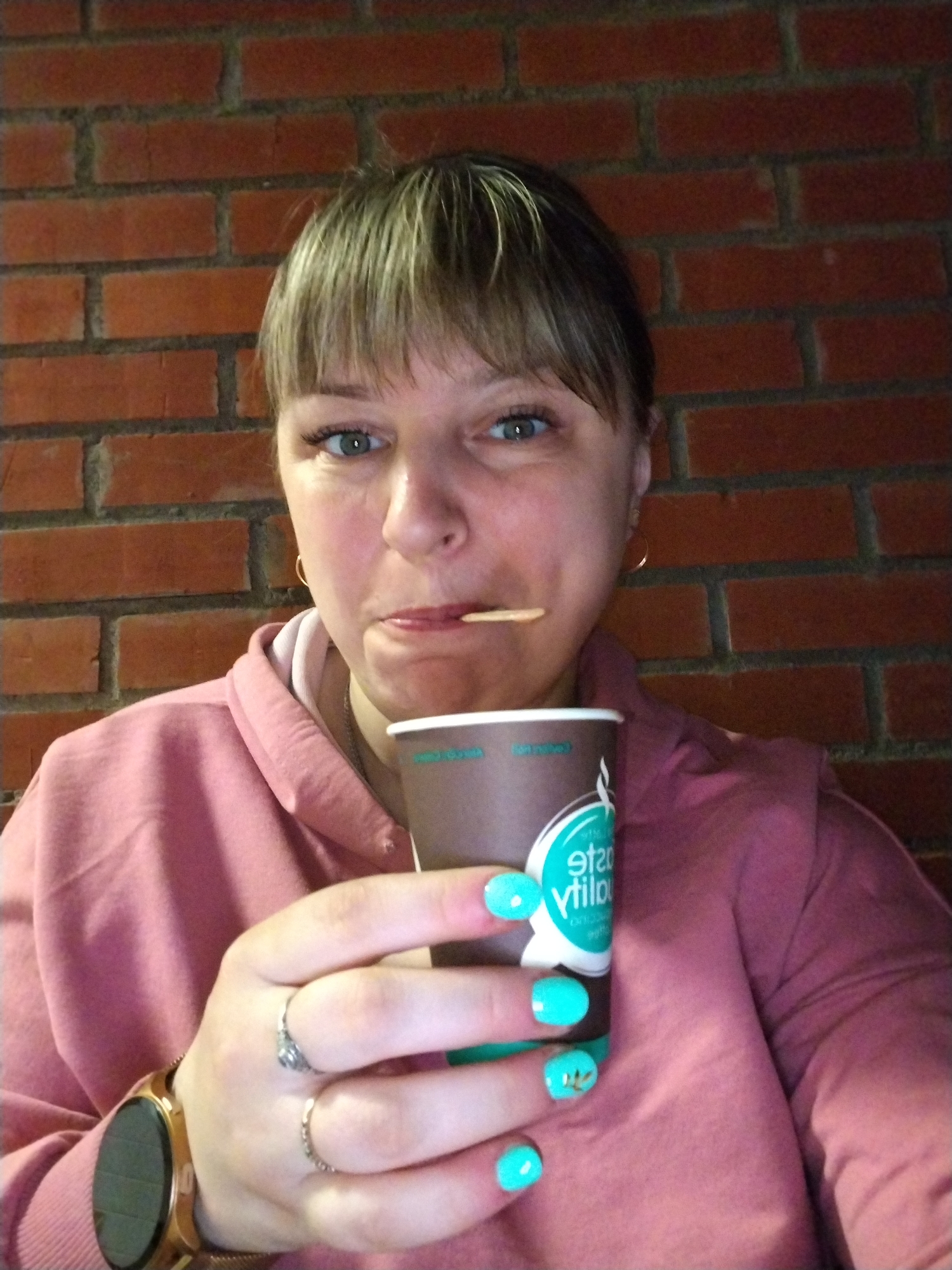 A woman is depicted in a close-up, chest-up portrait against a red brick wall, suggesting an indoor or sheltered setting in Шебунино, Russia. She has light brown hair with bangs, blue-green eyes, and a playful expression, accentuated by a wooden toothpick held between her lips. She wears a pink hooded sweatshirt, gold hoop earrings, a smartwatch with a gold-toned mesh band on her left wrist, and two rings on her left hand. Her fingernails are painted a bright turquoise, with a delicate gold leaf design visible on the ring finger. She holds a dark brown paper cup in her right hand. The cup features a white and teal logo with a stylized design, and the text "state quality" (visible in reverse as "ytilauQ etats"). The lighting suggests an indoor environment. The woman's direct gaze and the toothpick add a casual, slightly whimsical touch to the image.