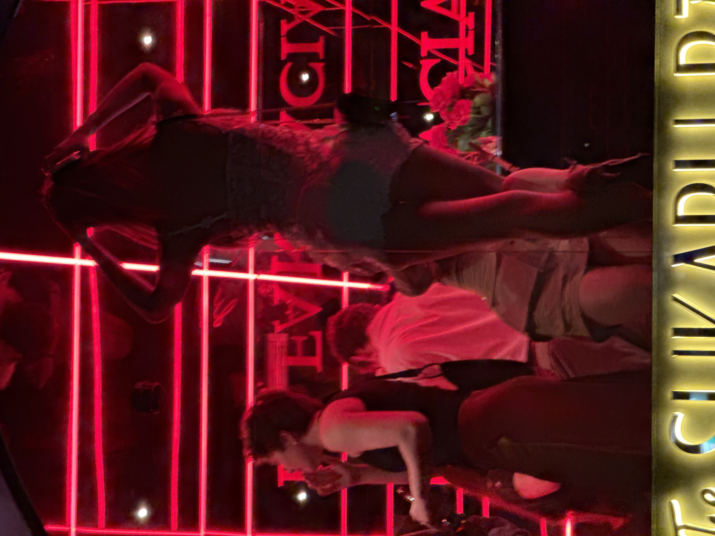 The image depicts a crowded, dimly lit nightlife venue in Milan, Italy, identified by partial signage as "The Clik" or "Clik ADULT" with additional background text "EVICE CLIX GIA". The scene is dominated by a grid-like pattern of intense red neon or LED strip lighting against a dark background.
In the foreground, a silhouetted female figure stands prominently on an elevated surface, possibly a stage or platform, with both arms raised above her head, suggesting dancing or posing. She is wearing a light-colored garment. To her left, another female individual is seen in profile, with dark hair, wearing a dark top, holding a small object to her mouth, possibly a cigarette or a drink. A male individual in a light-colored shirt is partially visible behind her. Floral or plant decorations, illuminated in red, are present on the right side of the background. The low light and specific signage indicate an indoor club or disco environment.