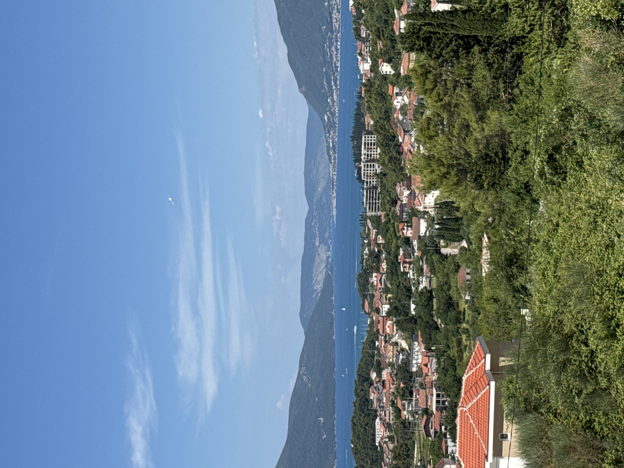 This is a landscape photograph taken from a high vantage point overlooking a coastal town and a bay. The sky is clear blue with a few wispy white clouds and a distant bird or aircraft. The foreground is dominated by lush green foliage, with a red-tiled roof of a building partially visible on the lower left. In the middle ground, a densely populated town with numerous buildings, primarily with red tile roofs, stretches across the landscape. Numerous trees are interspersed among the houses. Further in the distance, a wide body of blue water, likely a bay or sea, is visible, with several sailboats and what appear to be speedboats. Along the far edge of the water, mountains rise, their slopes covered in green vegetation. The overall scene depicts a sunny day in a Mediterranean or coastal region. Based on the context provided, this view is likely of Kavač, Montenegro, a settlement located near Tivat in the Bay of Kotor.