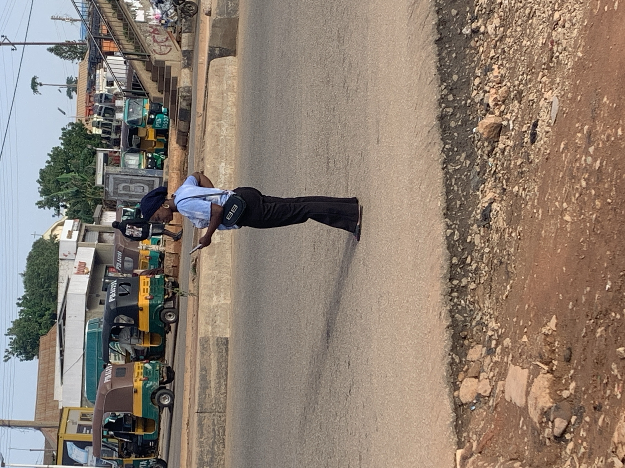 A woman in a blue t-shirt and black pants, wearing a dark headscarf, stands on an asphalt road, holding a smartphone. She has a black handbag with the letters "BB" on it slung across her body. In the background, several auto-rickshaws, also known as "Keke NAPEP" in Nigeria, are parked or in motion. Some of the auto-rickshaws have "PTA" and numbers on them. Buildings with signage, including what appears to be "BOJO" are visible behind the vehicles. A pedestrian overpass with stairs is also present. The ground in the foreground is unpaved and rocky. The scene is set in Gura Top, Nigeria.