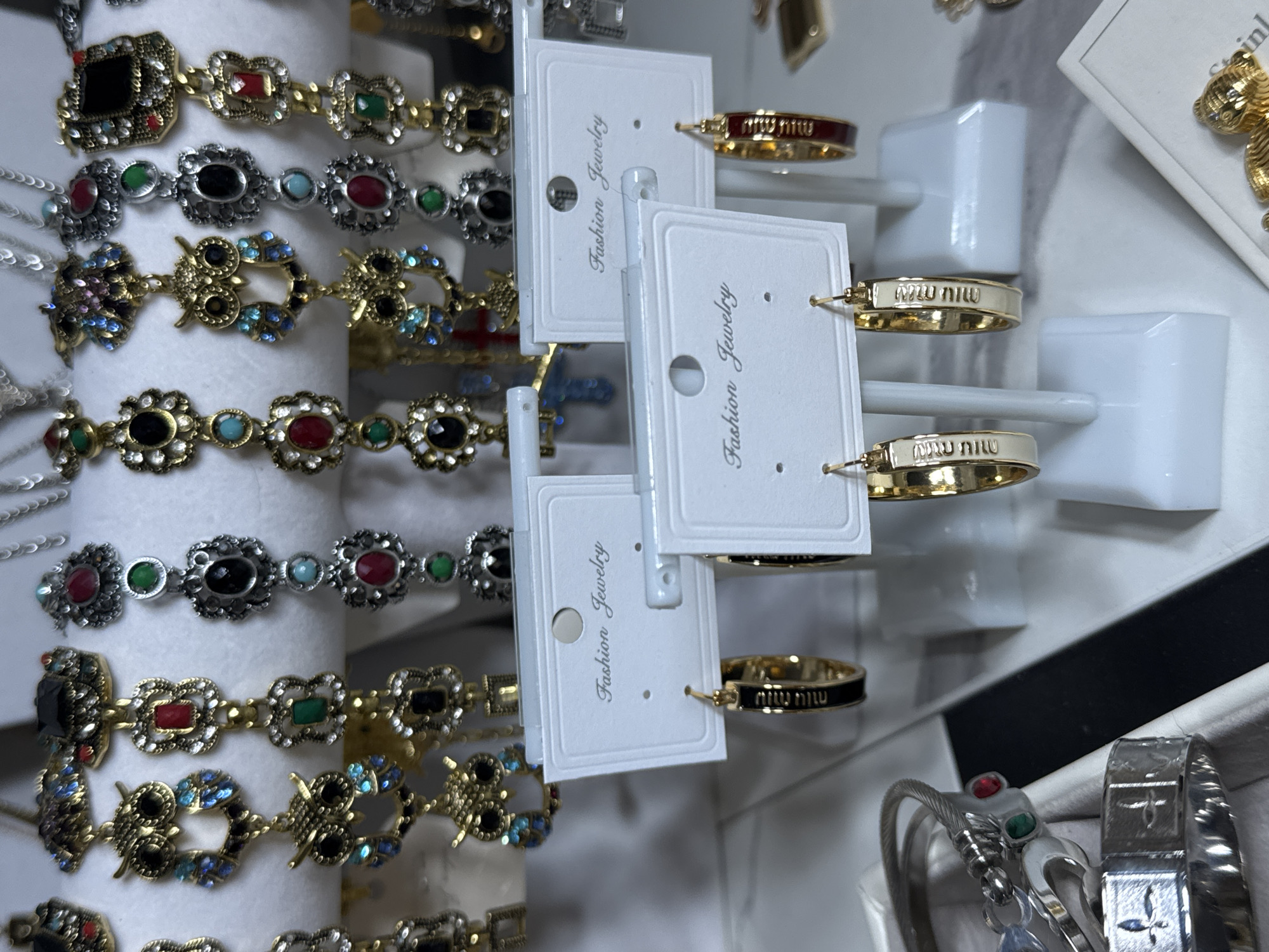 The image shows a close-up, eye-level view of several pieces of costume jewelry displayed on white cardstock tags and white plastic stands. The jewelry includes earrings, bracelets, and rings, featuring various embellishments such as colorful gemstones, owls, and ornate metalwork. The earrings in the foreground are hoop earrings with gold-colored metal and either black or red enamel, some with visible brand text. Behind these, rows of bracelets and other dangling earrings with intricate designs are visible. The overall setting appears to be a retail display, possibly in a jewelry store or market stall. The lighting is bright and even, highlighting the details of the jewelry. The background is blurred, suggesting a shallow depth of field. There is no indication of the location in the city of Wheeling, United States of America.