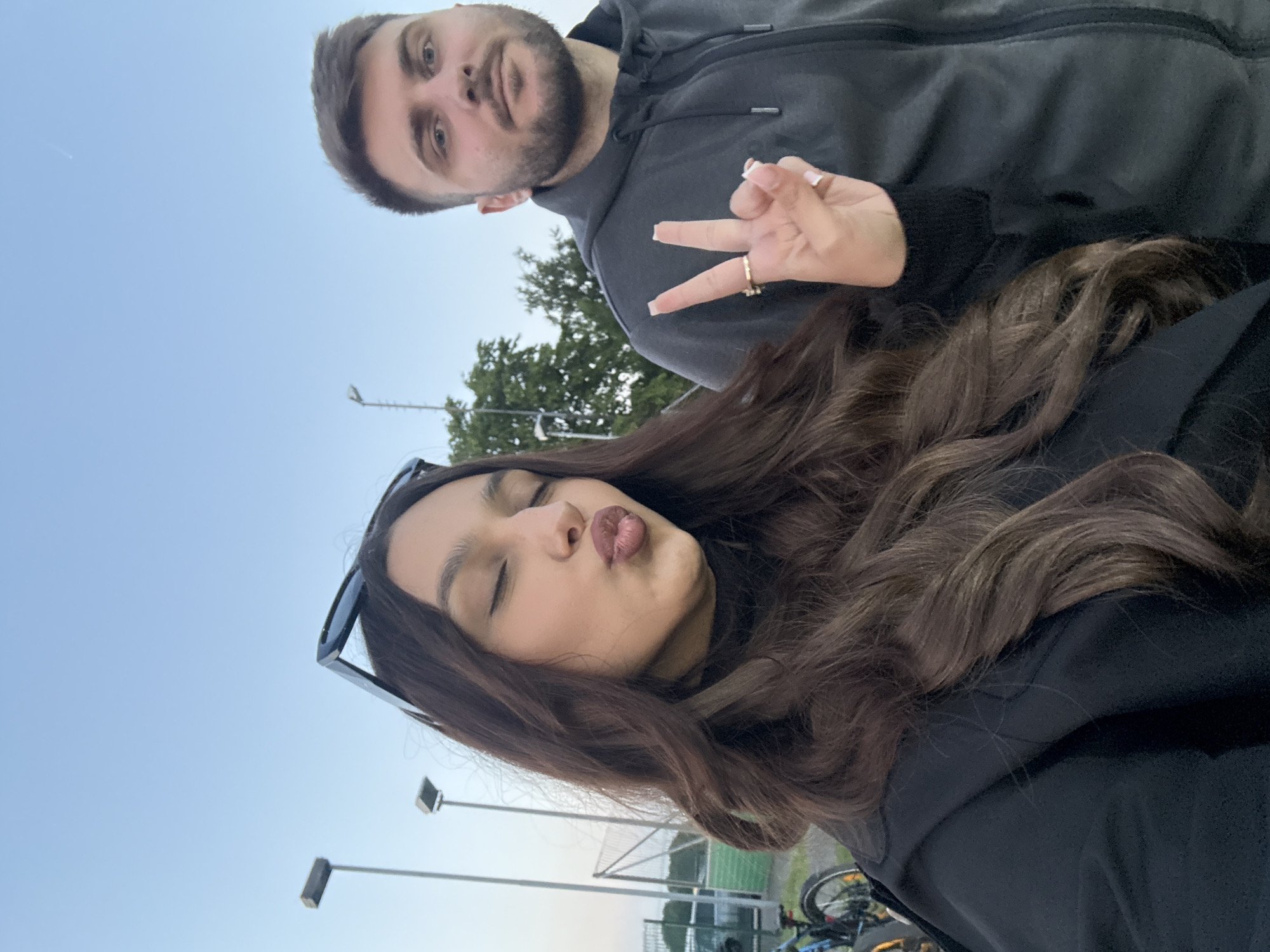 A medium shot taken outdoors shows a man and a woman. The woman is in the foreground, angled slightly to the right, and making a duck face. She has long, wavy brown hair and is wearing sunglasses pushed up on her forehead and a black jacket. The man is in the background, angled to the left. He has short dark hair, a beard, and is wearing a gray hoodie. The woman's hand is visible making a peace sign, with a gold ring on her finger. Street lights are visible in the background along with a soccer net and bicycles. The sky is a pale blue and suggests this may be a late afternoon or early evening photo. The location is Wegberg, Germany.