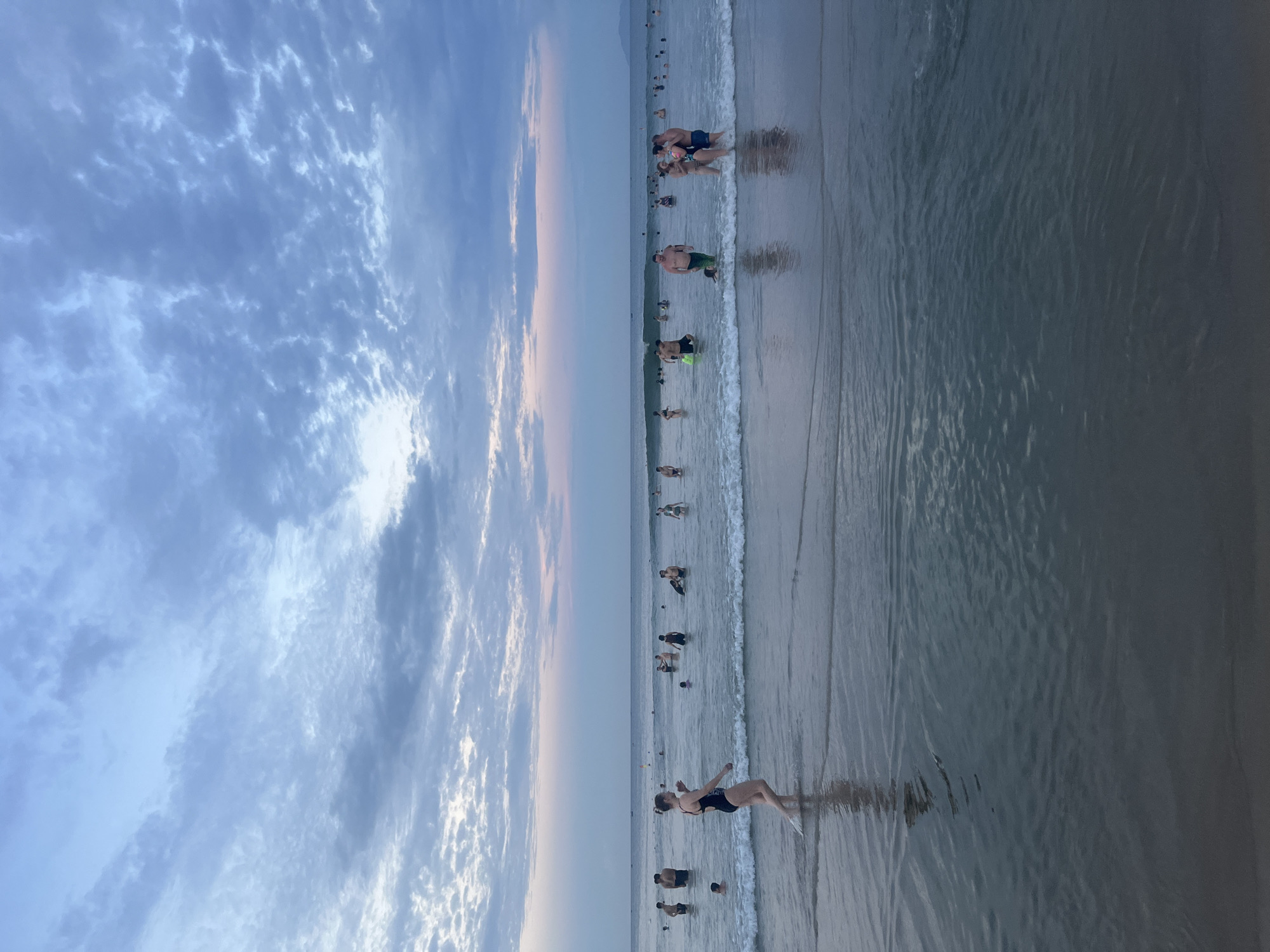 This image depicts a scene at My Khe Beach in Đà Nẵng, Vietnam, during what appears to be late afternoon or early evening. The sky is filled with a mixture of light and dark blue clouds, with a hint of pink on the horizon where the sun is setting. The ocean is calm with small waves rolling onto the shore. Numerous people are enjoying the water, some wading, others swimming. In the foreground, a woman in a black swimsuit is walking through the shallow water. Further out, groups of people are scattered across the sea. The overall atmosphere is relaxed and recreational, indicative of a popular beach destination.