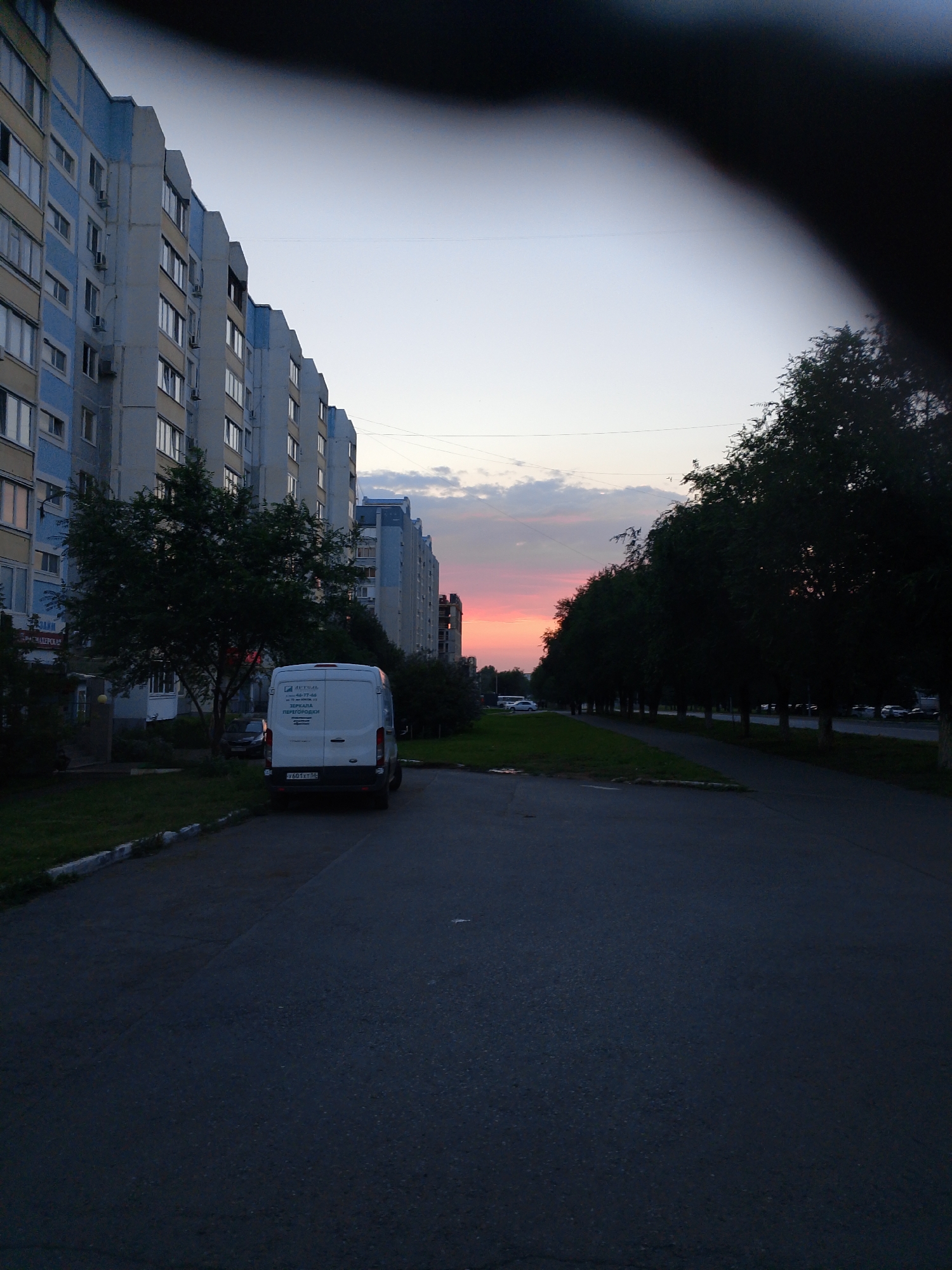 A residential street is depicted at dusk, featuring a multi-story apartment building on the left and a line of trees on the right. A white van is parked on the asphalt road in the foreground, facing away from the camera. The sky above is a pale blue, transitioning to hues of pink and orange near the horizon, indicating sunset. Power lines are visible crisscrossing the sky. The environment appears to be a typical urban or suburban setting.