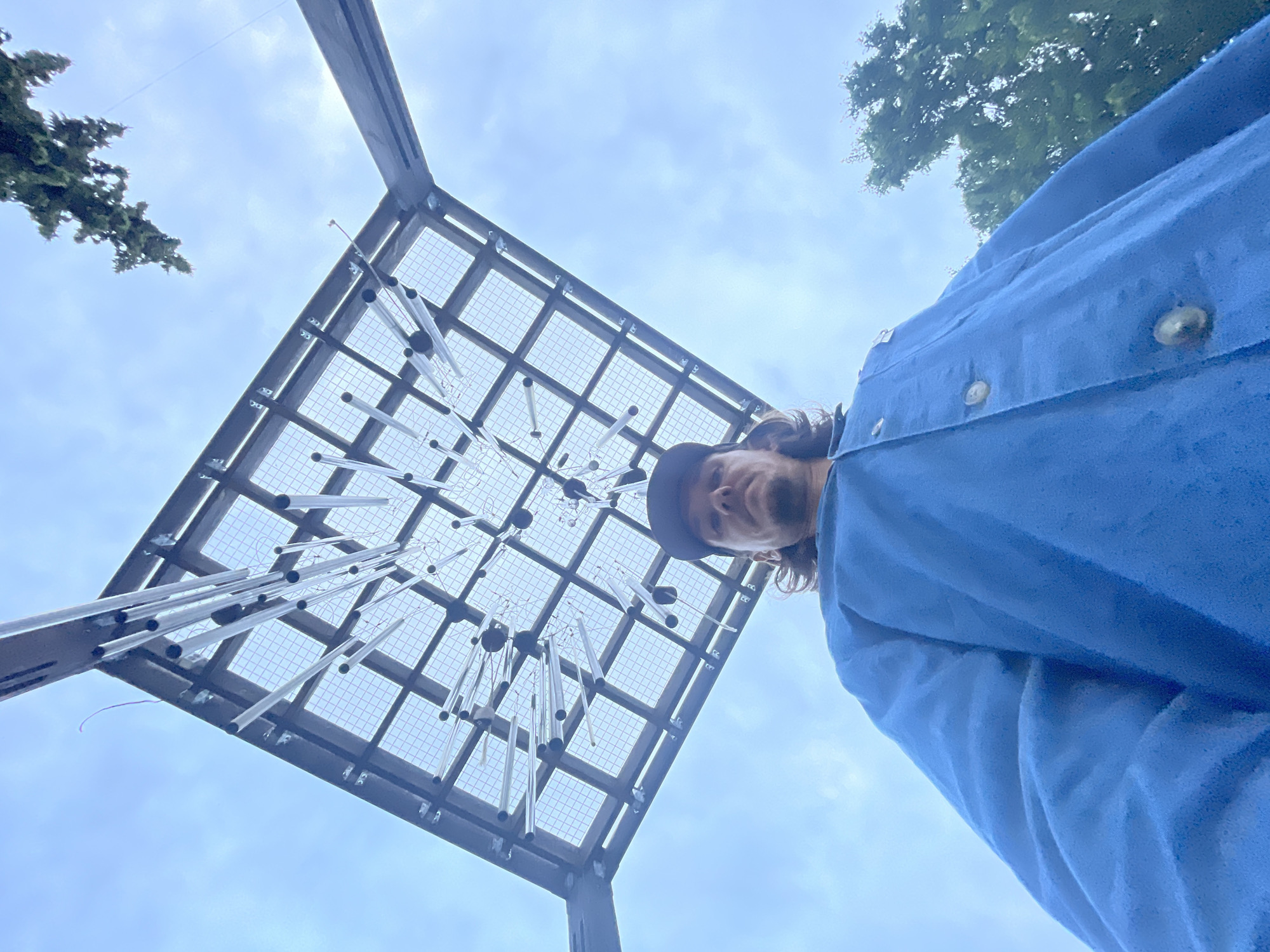 A low-angle, upward-facing photograph captures a single individual standing beneath a large, square overhead structure in the city of Unknown, Unknown, under a cloudy sky. The person, positioned in the lower-middle of the frame, is seen from the chest up, looking directly at the camera. They wear a dark baseball cap reversed, covering their long, dark brown hair. Facial features include a mustache. They are dressed in a medium-blue, long-sleeved shirt or jacket with several light-colored buttons visible on the front.
The prominent overhead structure, occupying the upper two-thirds of the image, is composed of dark metal beams forming a square grid. Within this grid, a finer mesh or net is visible. Numerous silver-colored cylindrical objects, possibly chimes or decorative tubes, varying in length and featuring dark caps, hang suspended at different heights and angles from the structure. The background consists of an overcast sky with light blue and gray clouds. Green foliage from trees is partially visible in the upper-right and lower-right corners of the frame. The lighting is diffused, indicating an overcast day.