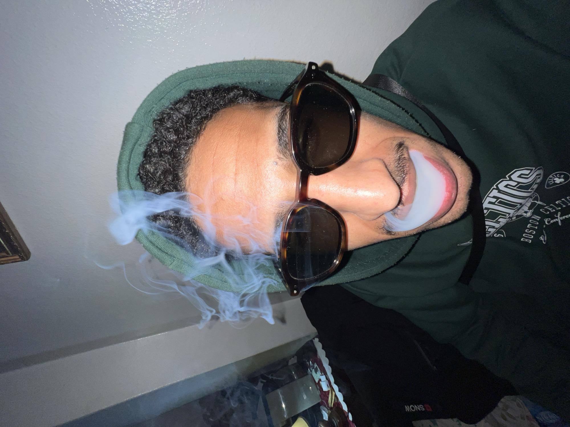 A person wearing sunglasses and a hooded sweatshirt is exhaling smoke. The sweatshirt appears to be green and bears the words "BOSTON CELTICS." Part of a picture frame is visible in the upper left corner of the frame. Below the person, there is what looks like a cluttered table with a black garment on top. The location is indoors with a white ceiling. There is no evidence of Tanger طنجة, Morocco.