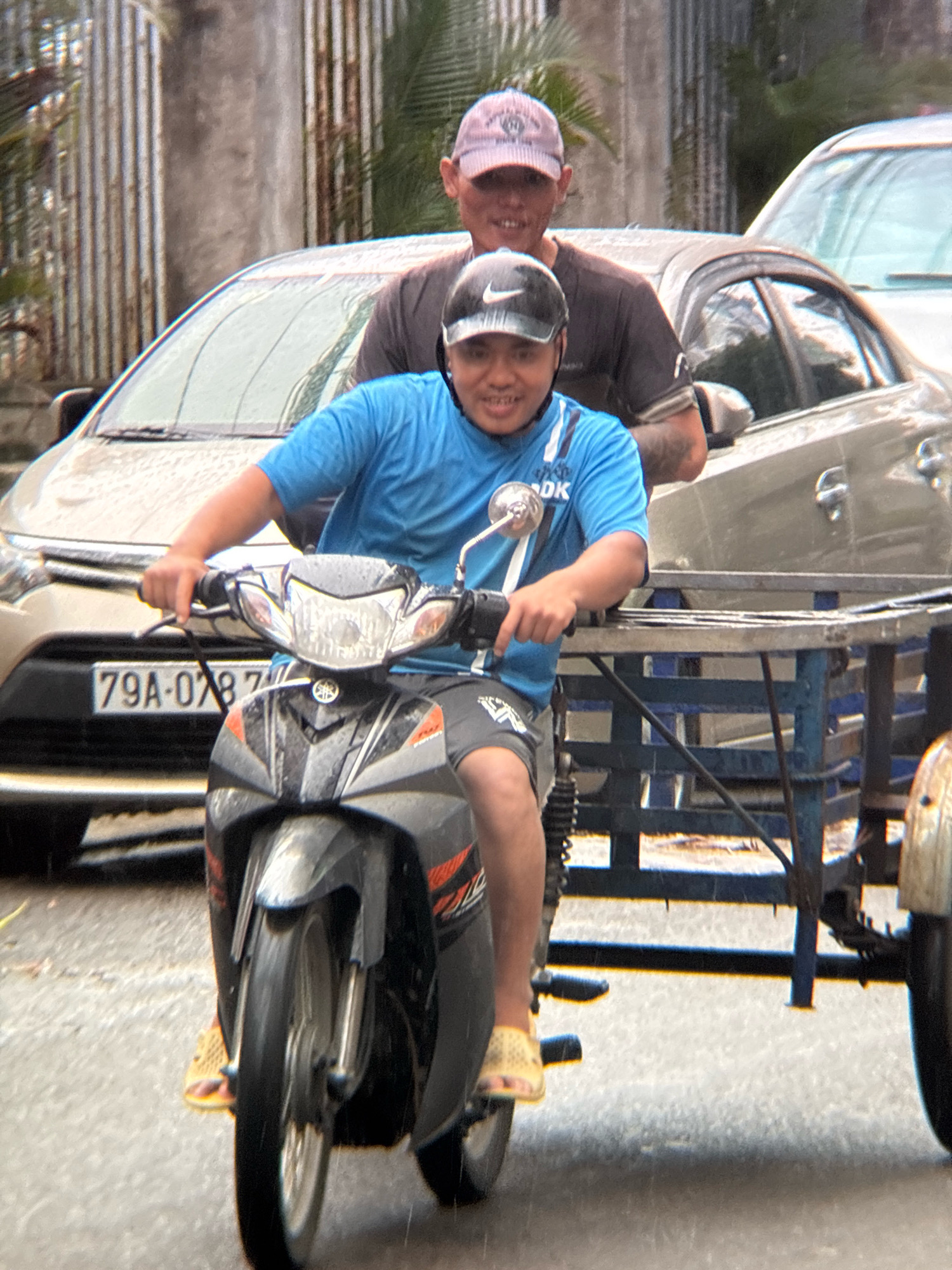 Two men are riding a motorcycle with a trailer in the city of Nha Trang, Vietnam. The man in front is driving the motorcycle and wearing a blue t-shirt and gray shorts. He is wearing a black helmet with a white Nike logo. The man behind him is wearing a dark t-shirt and a light-colored baseball cap. He is partially obscured by the man in front. The motorcycle has a license plate that reads "79A-078.77". The trailer is a blue metal frame with a black net. The motorcycle is in motion on a wet street, suggesting it may be raining. Cars are visible in the background, and there are metal fences with palm trees behind them.