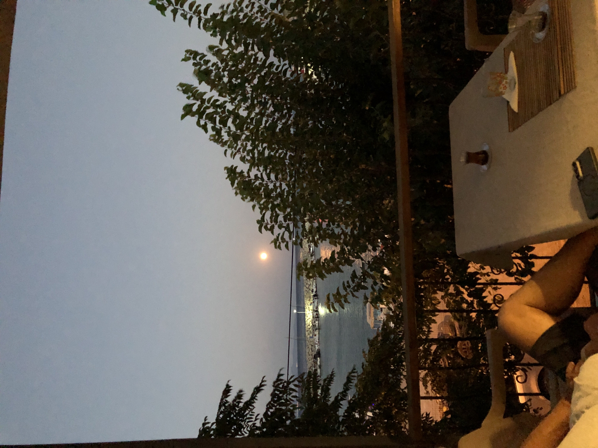 The image shows a balcony overlooking a harbor at dusk in Karaburun, Turkey. The full moon is visible in the sky. On the balcony, a table is set with two glasses of tea and a placemat. A person is lying on a lounge chair, partially visible. Trees and foliage frame the view of the harbor, which contains several docked boats and a pier with lights. The overall scene suggests a relaxed evening atmosphere.
