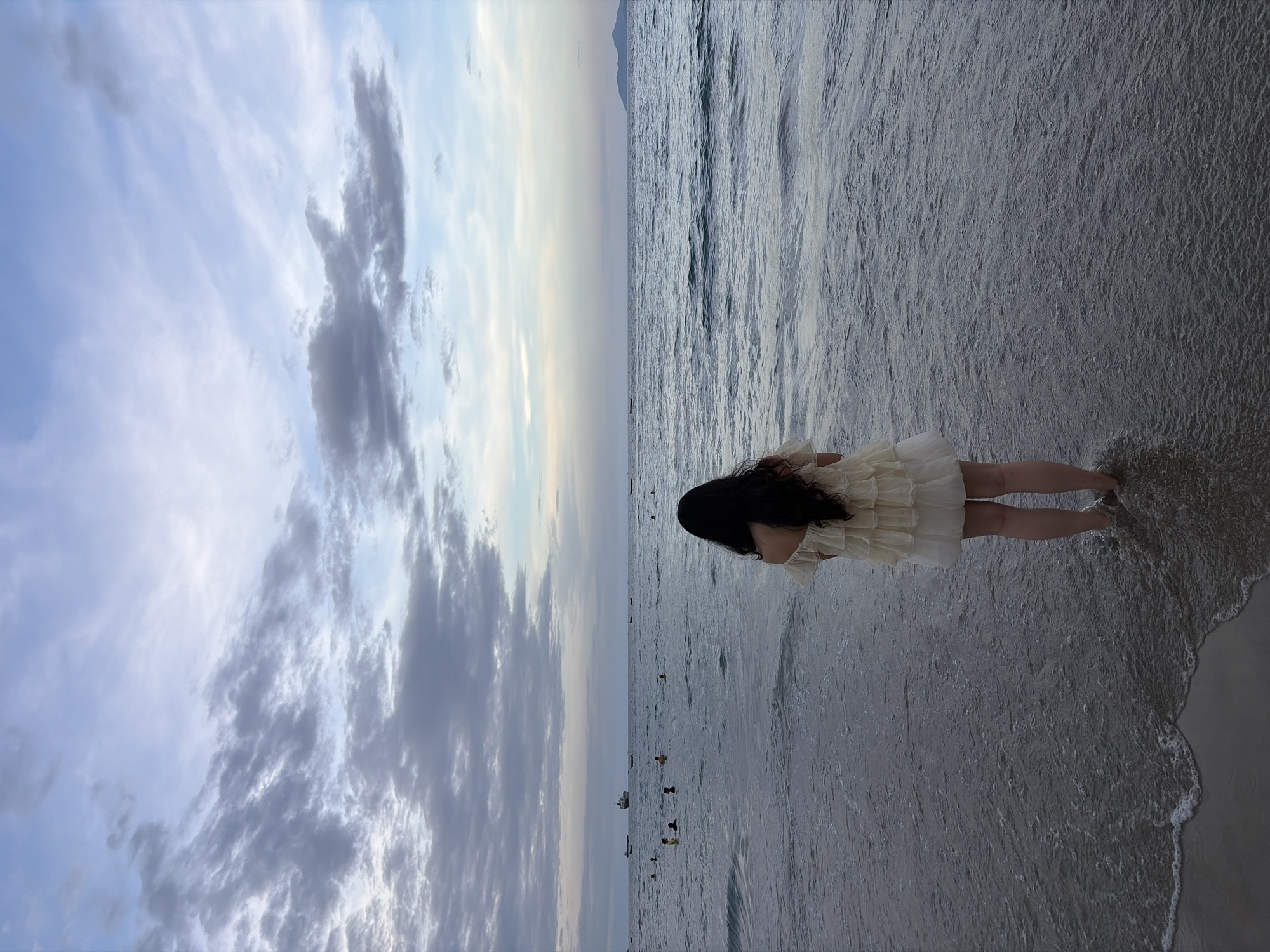 The image captures a serene moment on a beach in Đà Nẵng, Vietnam, likely during the late afternoon or early evening due to the soft, diffused light. The vast expanse of the ocean dominates the scene, with gentle waves lapping at the shore. The water is a deep blue-gray, reflecting the overcast sky above, which is adorned with streaks of white and gray clouds.
In the foreground, a young woman stands with her back to the camera, facing the sea. She has long, dark hair and is wearing a light-colored, ruffled dress. Her feet are submerged in the shallow water at the edge of the beach.
Further out in the ocean, several other people can be seen enjoying the water, some swimming and others wading. A few small boats are scattered across the horizon, and a larger ship is visible in the distance. To the far right of the frame, a mountainous silhouette of an island or coastline emerges from the haze. The overall atmosphere is one of peaceful contemplation and connection with nature.