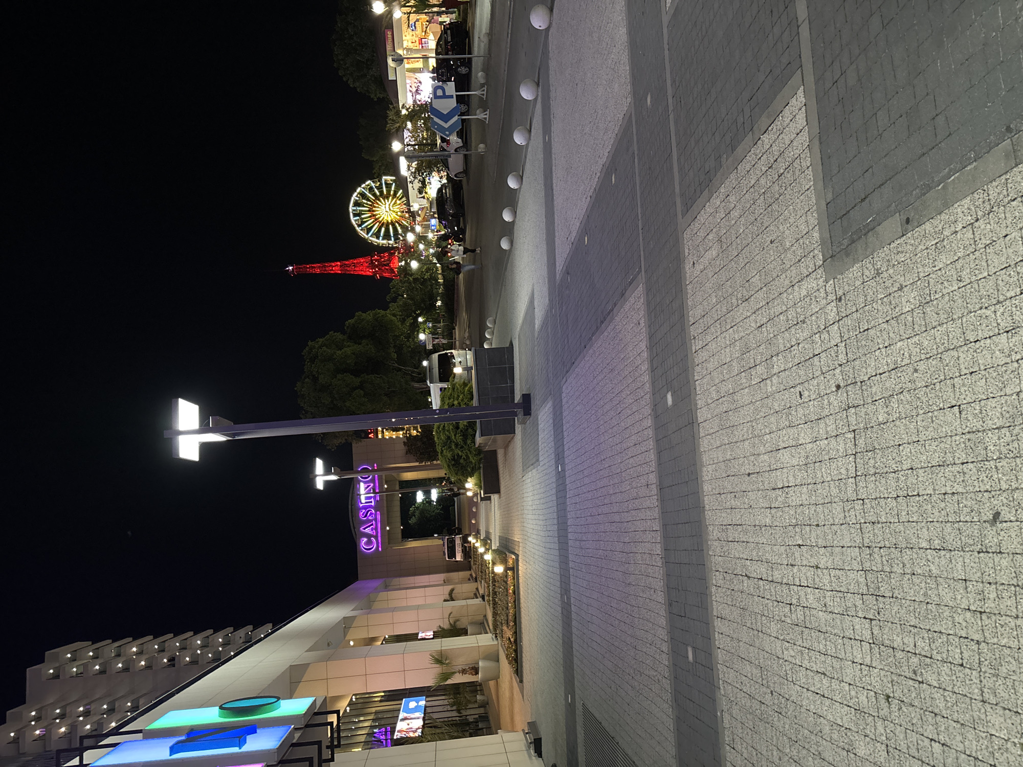The image depicts the entrance to a casino in Varna, Bulgaria, at night. A modern building with illuminated signage, including the word "CASINO" in large purple letters, is visible. The area in front of the casino features a paved walkway with decorative lighting and planters. In the background, a Ferris wheel and a tall, red illuminated tower, possibly a replica of the Eiffel Tower, suggest an amusement park or entertainment area. Several vehicles, including cars and a white van, are parked in the vicinity. Two individuals are walking in the middle ground, and a parking sign is also visible. The overall scene conveys a lively urban atmosphere at night, characteristic of a tourist or entertainment district.