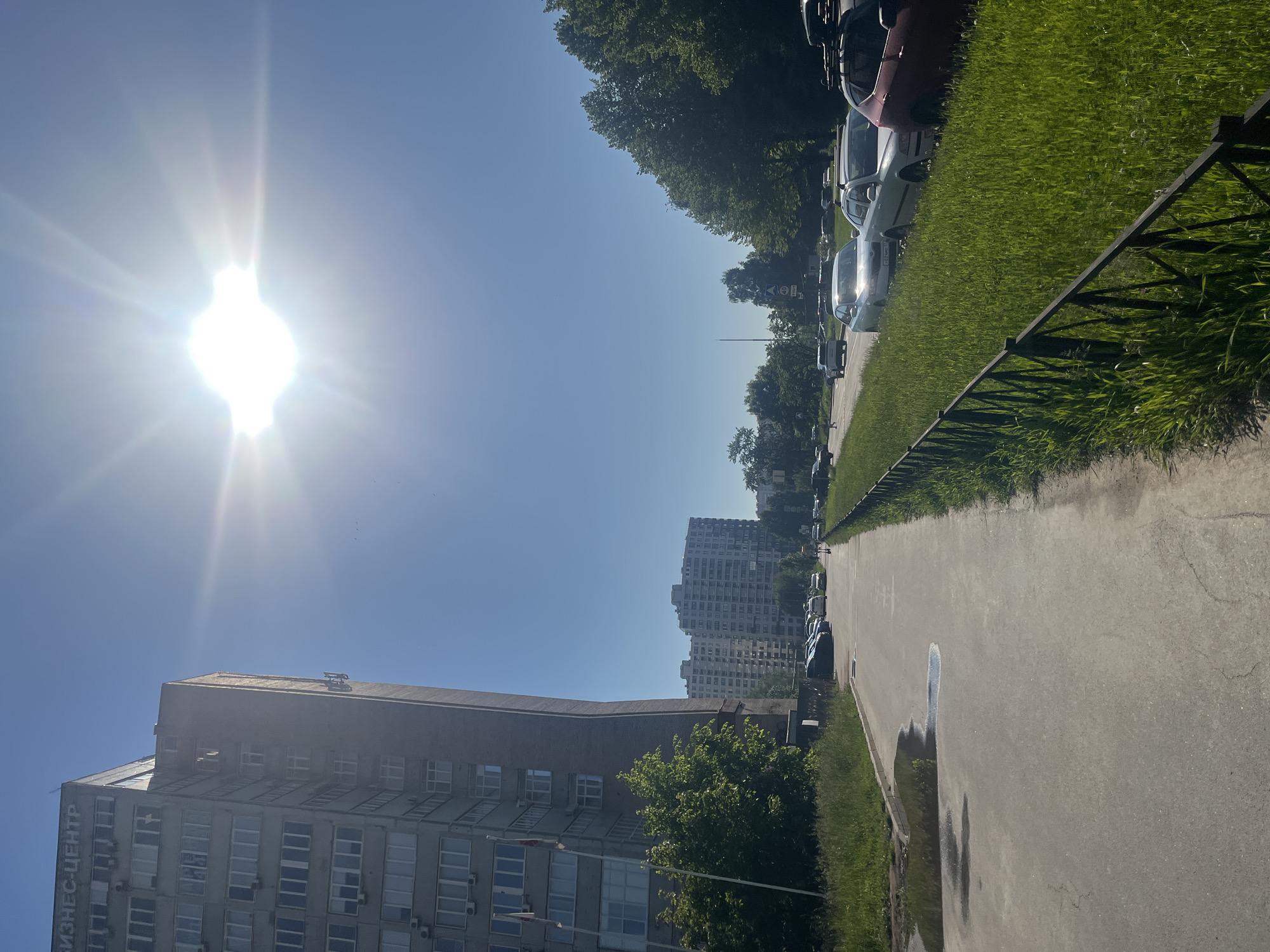 The image captures a bright, sunny daytime scene in what appears to be an urban or suburban area. The sun, high in the sky, emits a strong flare on the left side, indicating midday or early afternoon. The sky is clear and blue, devoid of clouds.
In the foreground, a large, multi-story building with numerous windows occupies the left side of the frame. Vertical Cyrillic text, "БИЗНЕС-ЦЕНТР" (BUSINESS-CENTER), is visible on its side. Further in the background, a taller, multi-storied residential-style building can be seen.
On the right, a paved road or path runs alongside a strip of vibrant green grass, separated by a dark metal fence. Several cars of various models and colors are parked along the edge of this green strip, partially obscured by lush trees that line the road. Further down the path, a large puddle of water is visible on the asphalt, reflecting the sky. The presence of Cyrillic text on the building suggests a location in a Slavic-speaking country. No specific activity or people are depicted; it is a still street scene on a clear, bright day.