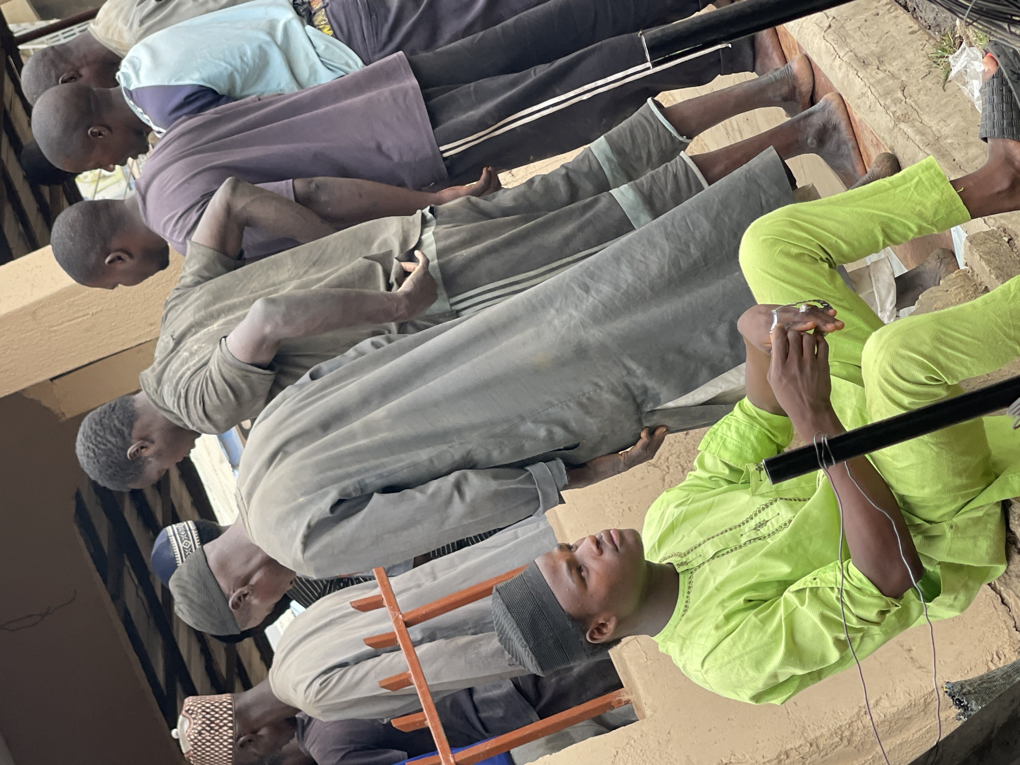 A group of at least seven men are engaged in prayer within what appears to be an outdoor setting in Zaria, Nigeria. The men are lined up in a row, kneeling. The foreground features a man in a lime green shirt and pants. Other men wear a variety of garments, including gray robes, purple shirts, and some with caps. In front of one of the men lies a ladder-like structure. The ground is light brown. There is a pole above the men.