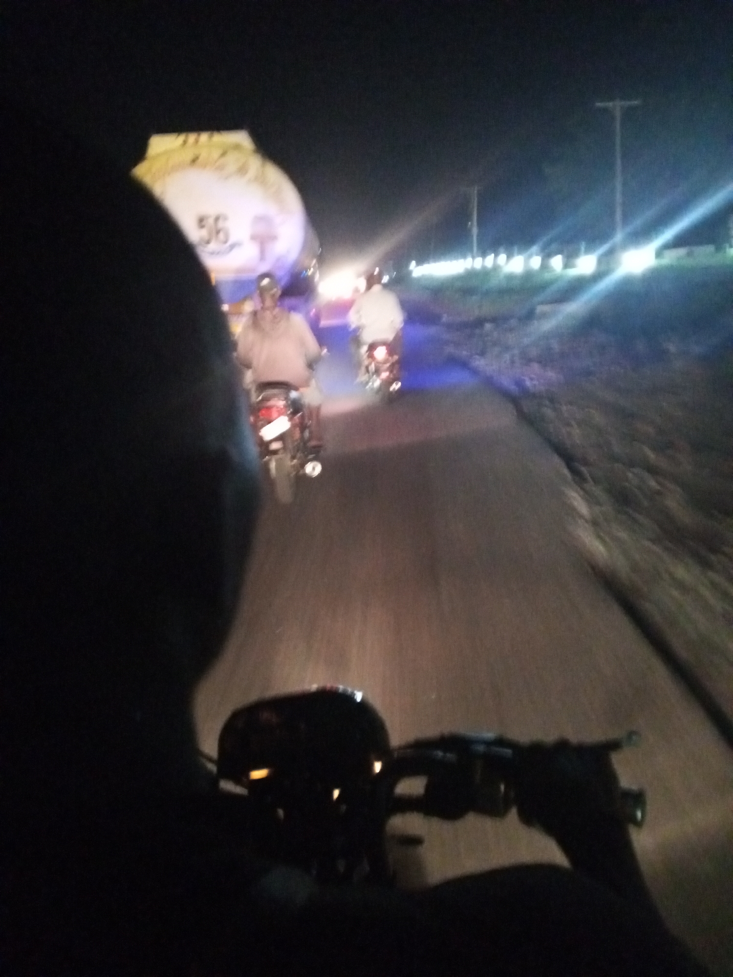 The image depicts a road in what appears to be a rural or semi-rural area at night. The photo is taken from the perspective of someone riding a motorcycle. Ahead, two other motorcycles are traveling in the same direction, their riders are visible. Further ahead, a large tanker truck is present on the road. Street lights line the side of the road in the distance. The image suggests the scene is on a road outside the city of Lokoja, Nigeria.