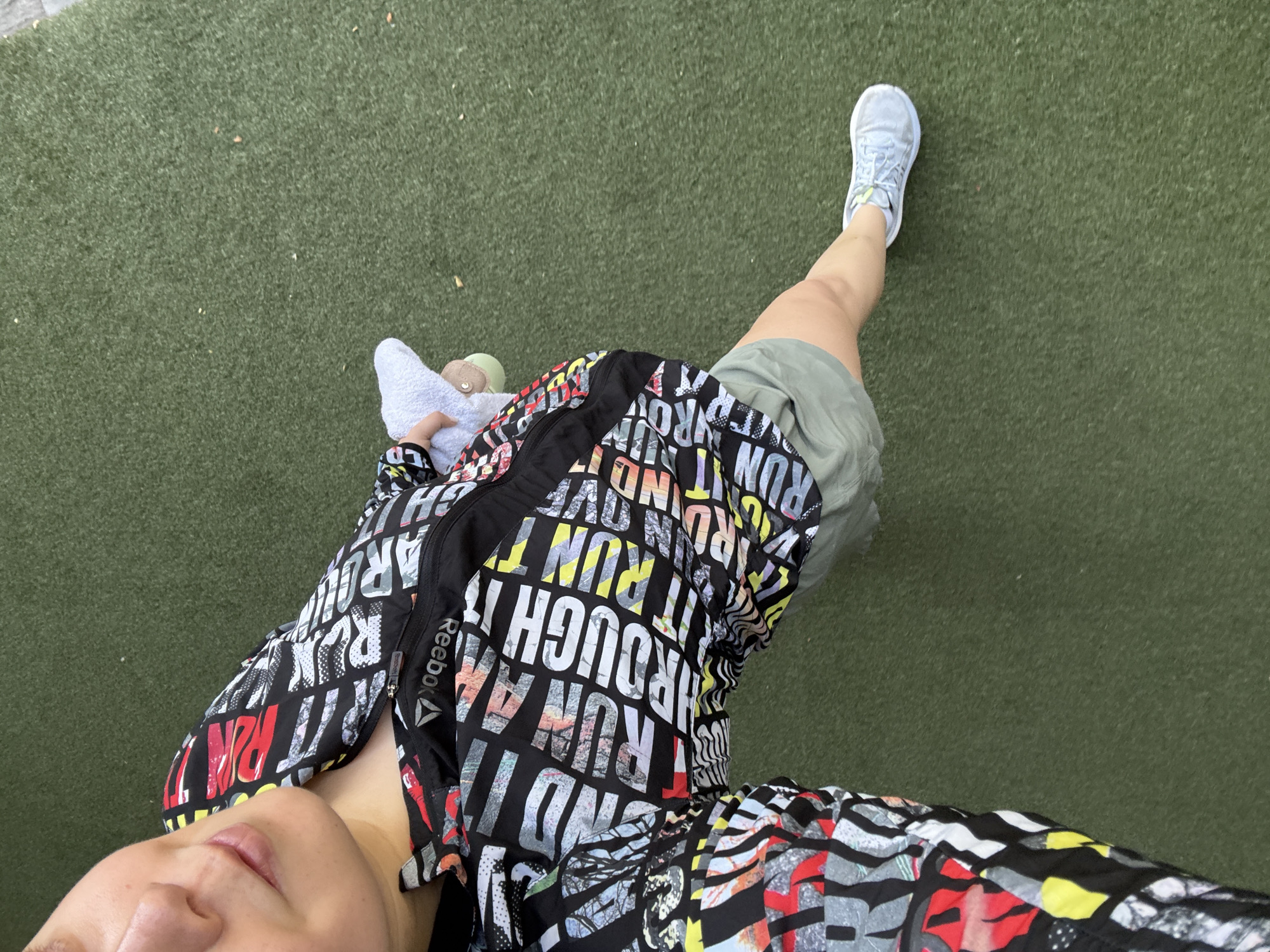 A first-person, high-angle shot shows an individual standing on a green surface, possibly artificial grass. The person is wearing a colorful Reebok jacket with the words "RUN" and "THROUGH" in large letters. The individual also has on gray shorts and light-colored sneakers. In the left hand, a white towel is held. Part of the person's face is visible, including the lips and nose. The location is not identifiable as being in Mexico City from the image alone.