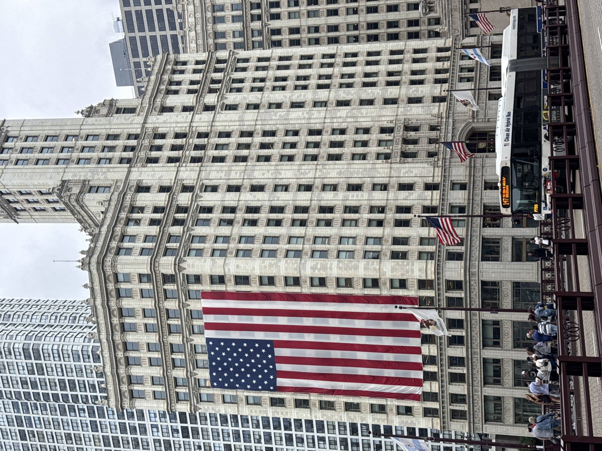 The image depicts the iconic Wrigley Building, a skyscraper located at 400 North Michigan Avenue in Chicago, Illinois, United States of America. A large American flag is prominently displayed, hanging down the side of the building. In front of the building, a clean air hybrid bus is seen passing by, displaying the route number 147. Several people are visible on a pedestrian bridge in the foreground, observing the scene. The sky is overcast, suggesting a cloudy day. The architecture of the Wrigley Building, with its intricate stone detailing and numerous windows, is characteristic of early 20th-century Beaux-Arts style. Adjacent to the Wrigley Building, other tall buildings can be seen, contributing to the urban cityscape of Chicago. Several flags, including the Illinois state flag and the flag of Chicago, are also visible on poles in front of the building.