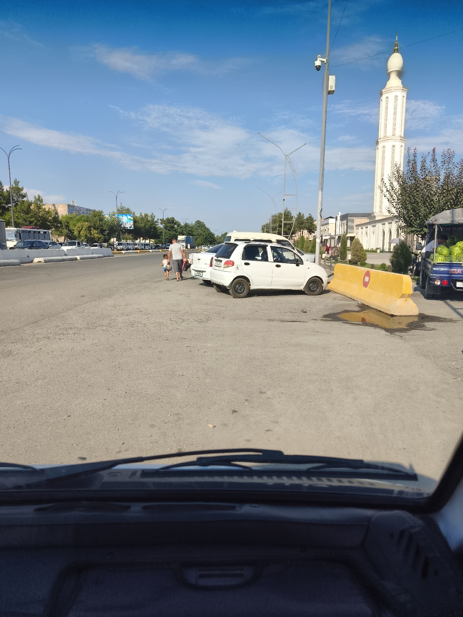 The image depicts an outdoor scene in Buz, Uzbekistan, on a clear day with a blue sky. In the foreground, the interior of a vehicle is visible, including the dashboard and windshield wipers. The road in front of the vehicle is paved with asphalt and shows some wear and tear, with a puddle of water and some debris present.
Several vehicles are parked or driving on the road. Two white cars are prominent, one of which appears to be parked somewhat haphazardly near a large, yellow concrete barrier. A blue truck with a canopy, loaded with watermelons, is parked on the right side of the road. A man and a child are walking away from the camera on the left side of the road, heading towards the white cars.
In the background, a tall, ornate minaret of a mosque stands against the sky, indicating a religious or cultural landmark. Trees line the sides of the road, and other buildings and vehicles are visible in the distance. A security camera is mounted on a pole near the minaret. The overall impression is a street scene in a city, with everyday activities taking place.