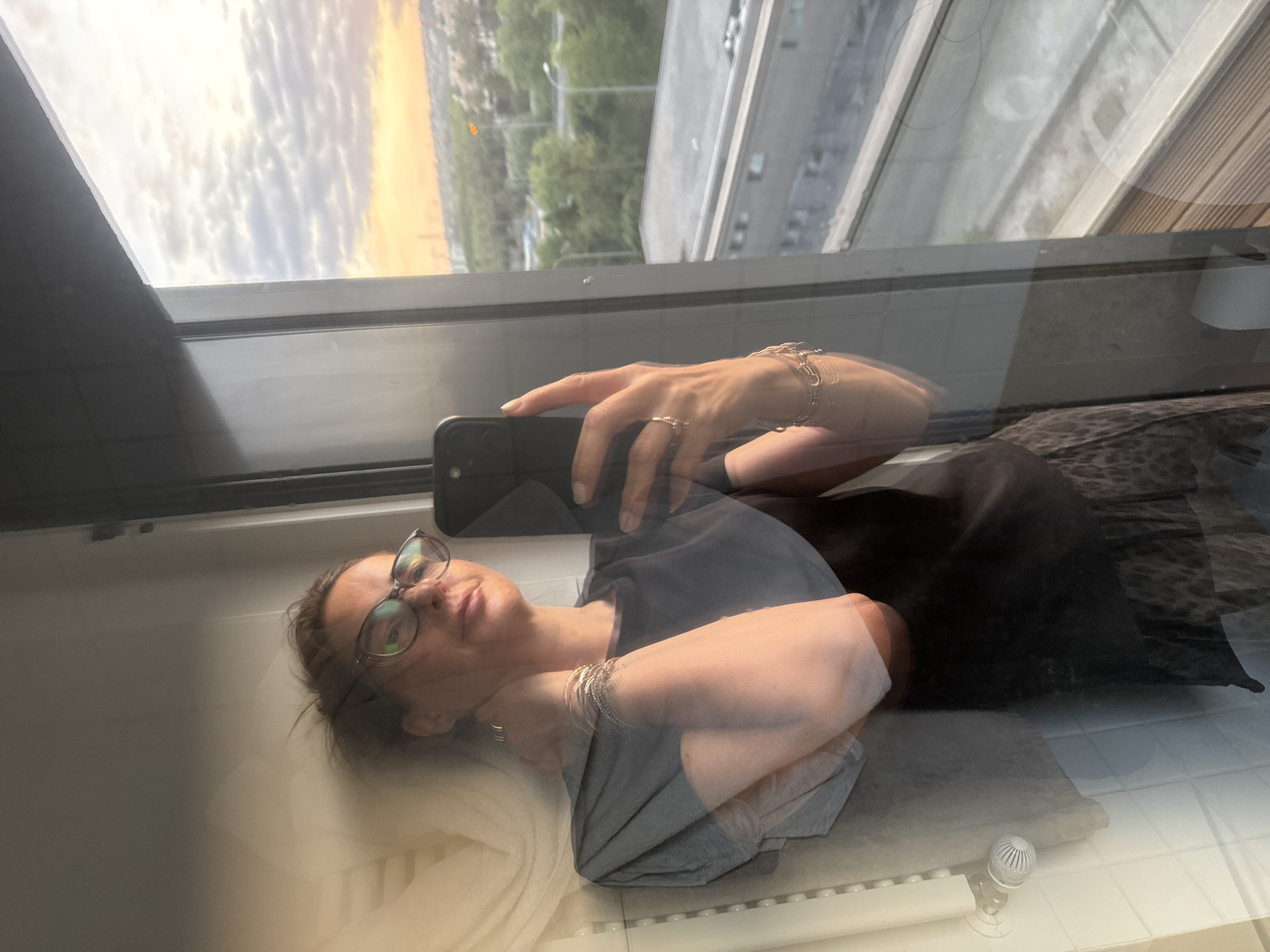 A woman wearing glasses and jewelry lies on a surface near a window, taking a selfie with her smartphone. The window reflects a cityscape, seemingly taken at sunset or sunrise. The location appears to be in Loures, Portugal, based on visible urban elements in the landscape.
