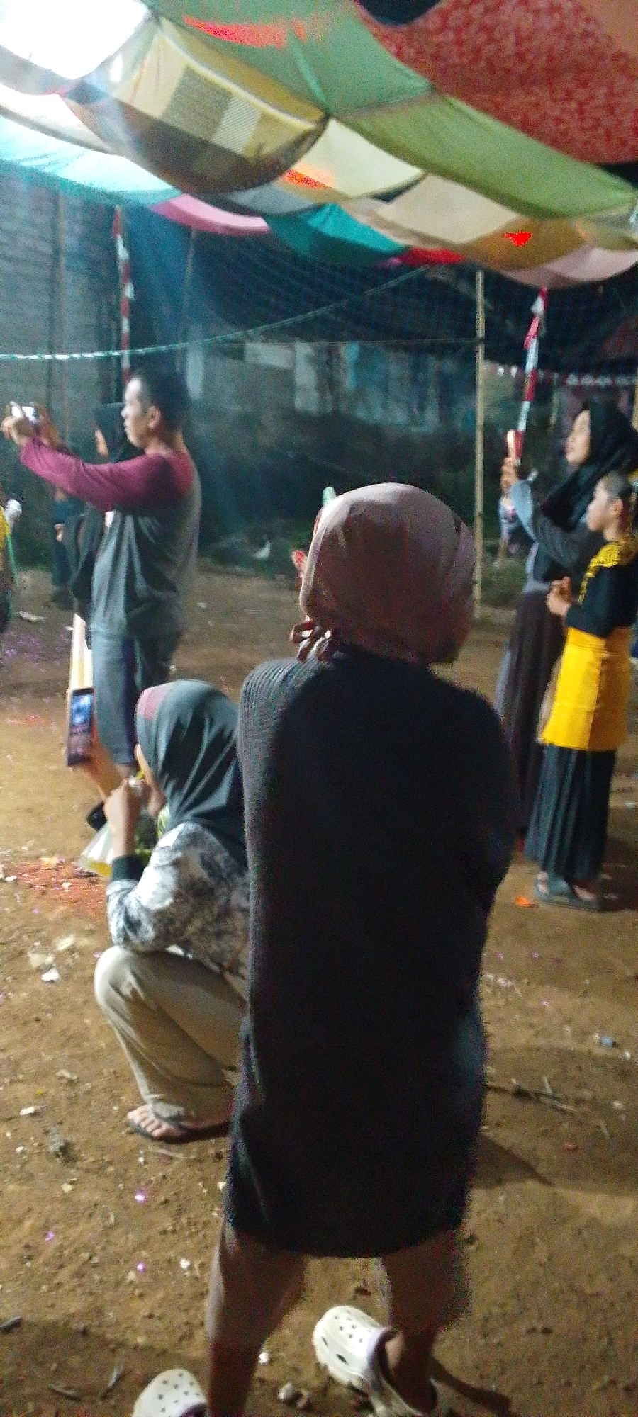 The image depicts an outdoor gathering at night, likely in Cilaku, Indonesia, given the context. Several individuals are visible, some engaged with personal devices. In the foreground, a person wearing a dark, long-sleeved top and a light-colored headscarf is viewed from behind, facing away from the camera. To the left, another person in a grey long-sleeved shirt and dark pants holds up a mobile device, seemingly taking a photo or video. Further left, a person wearing a hijab and a patterned shirt is crouched down, also looking at something on the ground. In the background, a person in a black hijab and another in a black garment with a bright yellow vest are standing. The ground appears to be dirt and debris. Above, a canopy made of fabric panels in various colors like green, white, and red is suspended, suggesting an event or makeshift shelter. The overall atmosphere appears casual, possibly a community event or celebration.