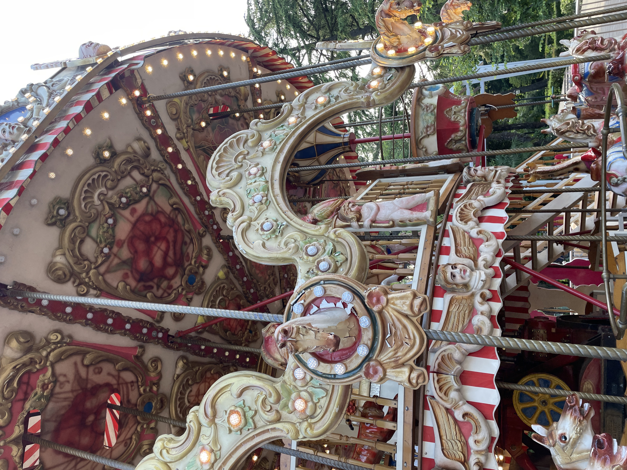 This is a close-up, eye-level, outdoor, daylight shot of a brightly lit, ornate carousel. The carousel features elaborate gold-colored carvings, red and white striped awnings, and various figures, including horses, cherubs, and a bird. The carousel is decorated with many small, warm-toned lights. In the background, lush green trees and foliage are visible, suggesting a park or garden setting. The overall impression is one of festive and vintage amusement. The location is identified as Sochi, Russia, though no specific landmarks within Sochi are visible in the image.