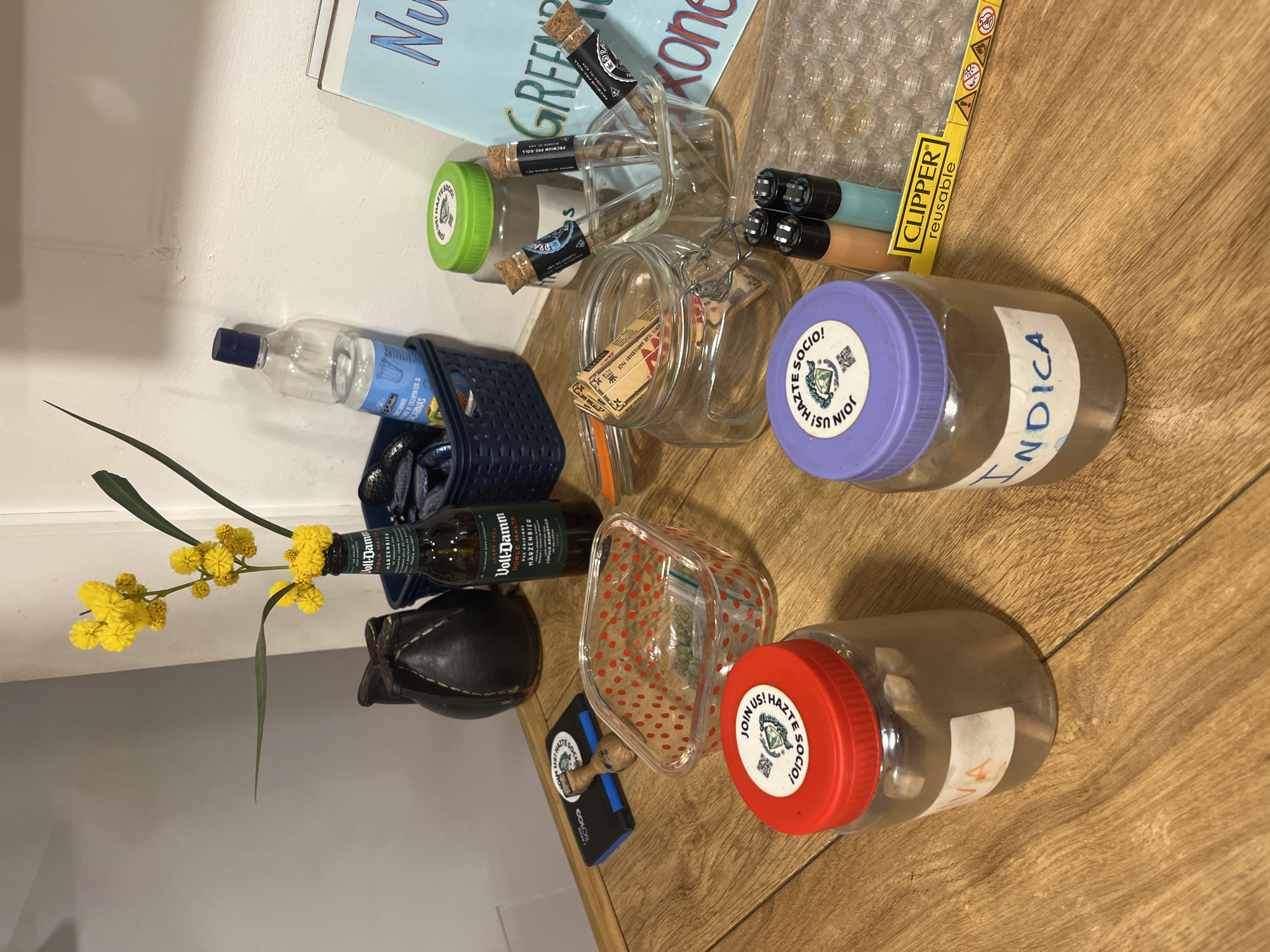 This image displays a collection of items arranged on a light wooden surface, likely a table or floor, next to a white wall. The scene suggests a personal, casual indoor setting.
Prominent objects include several clear jars and containers. One jar with a purple lid is labeled "INDICA" and has a sticker that reads "JOIN US! HAZTE SOCIO!". Another clear jar with a red lid also features the "JOIN US! HAZTE SOCIO!" sticker. Inside a clear, red polka-dotted plastic container, what appears to be cannabis is visible. Three small glass tubes, labeled "PREMIUM PRE-ROLL," contain pre-rolled items. A small box of "RAW ARTISAN ART OF ROLLING" papers and several "CLIPPER reusable" lighters are also present, further indicating items related to cannabis consumption. A wooden stamp with the "JOIN US! HAZTE SOCIO!" logo reinforces this theme, suggesting affiliation with a cannabis social club.
Other objects include a bottle of Voll-Damm Marzenbier (a Spanish beer brand), a small plastic bottle of water with partially visible text "RECORANTE" and "SICKLAMATOS & ACARINAS," a dark blue woven basket, and a small black leather-like pouch. A sprig of yellow flowers, resembling mimosa, adds a decorative touch. Partially visible books with titles "Nu," "GREE," and "Kon" are seen in the background.
The visible Spanish text ("Hazte Socio!", "INDICA," and the Voll-Damm beer) strongly suggests a location in a Spanish-speaking country, consistent with the provided context of Barcelona, Spain, where cannabis social clubs are common. No specific activity is taking place, but the arrangement of items implies preparation for or use of recreational cannabis. The time of day and weather are not discernible due to indoor lighting. No explicit emotions are conveyed.