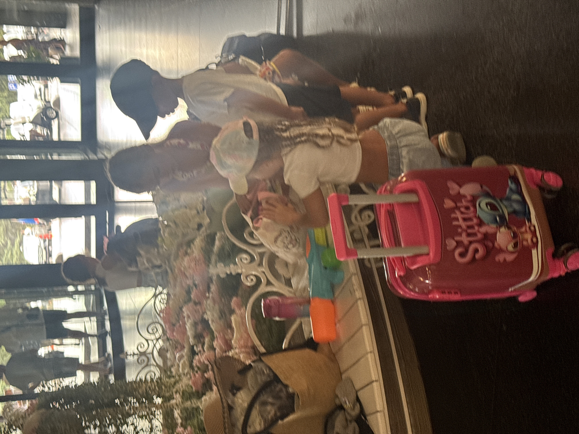 In an indoor, tiled environment, likely a hotel lobby, a group of children is gathered near a decorative bench. A young boy, wearing a black baseball cap and white t-shirt, stands to the right, facing left. To his left, a young girl with a pink and white tie-dye hat is looking down at something she is holding. Beside her, another girl, seen from the back, wears a white crop top and light blue shorts, her hair in braids. In the foreground, a bright pink, child-sized rolling suitcase decorated with Stitch characters is positioned on the dark floor. The bench behind the children is adorned with an abundance of pink and white artificial flowers. On the bench, a brown woven bag, a purple water bottle, and a child's toy gun are visible. In the background, through a glass-paneled entrance, other people are visible, along with what appears to be a golf cart and a street with cars. The lighting suggests it is daytime.