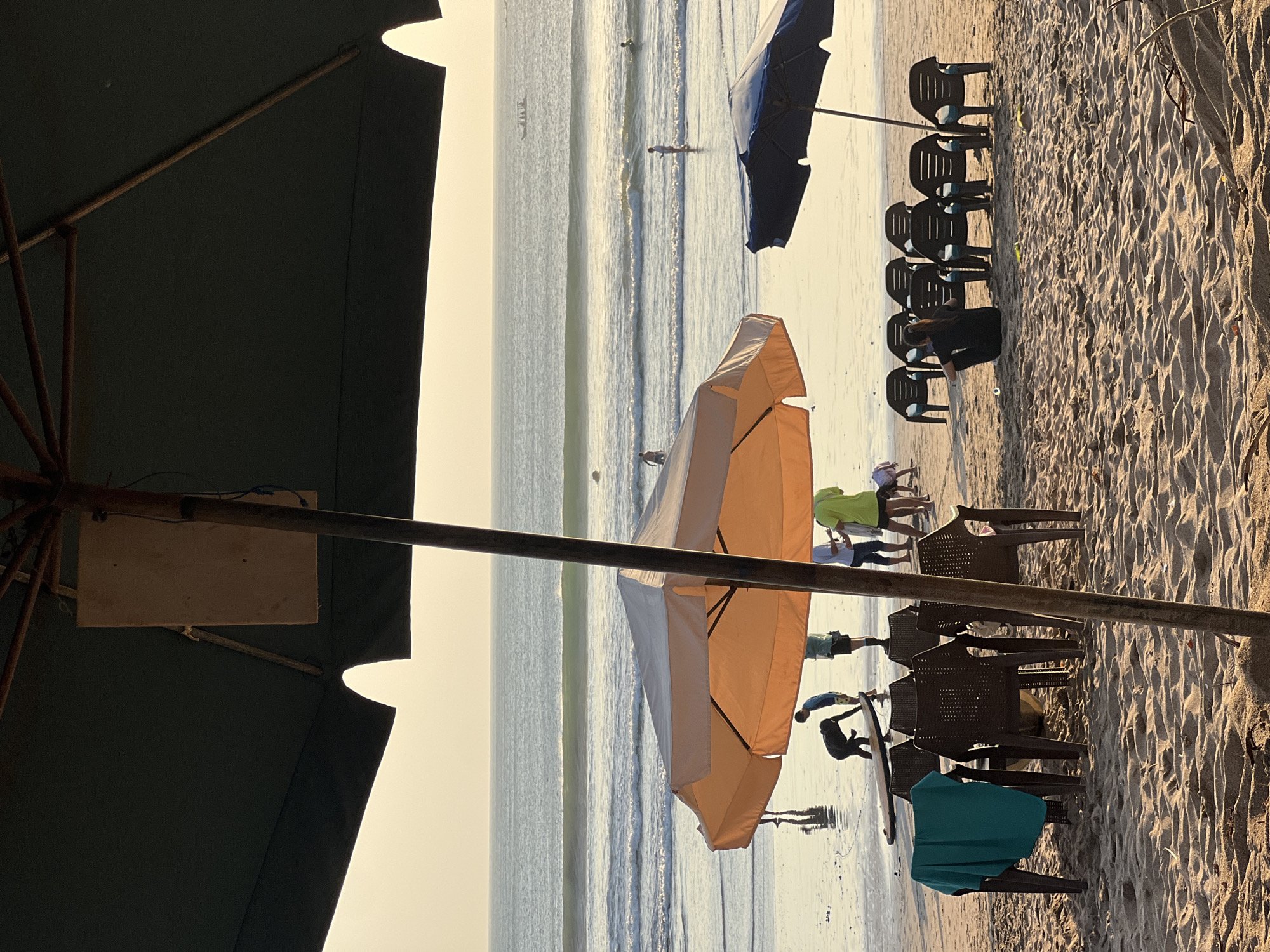 The image depicts a beach scene in Kuta, Bali, Indonesia, at sunset. In the foreground, two large beach umbrellas cast shadows on the sand, under which a group of plastic chairs are arranged. A teal towel is draped over one of the chairs. Further in the background, the ocean stretches out to the horizon, with gentle waves breaking on the shore. Several people are visible on the beach and in the water. One person is bent over near a surfboard, while others are walking along the water's edge or swimming. The sky is a warm, golden hue, indicating the setting sun. The scene conveys a relaxed and tranquil atmosphere.