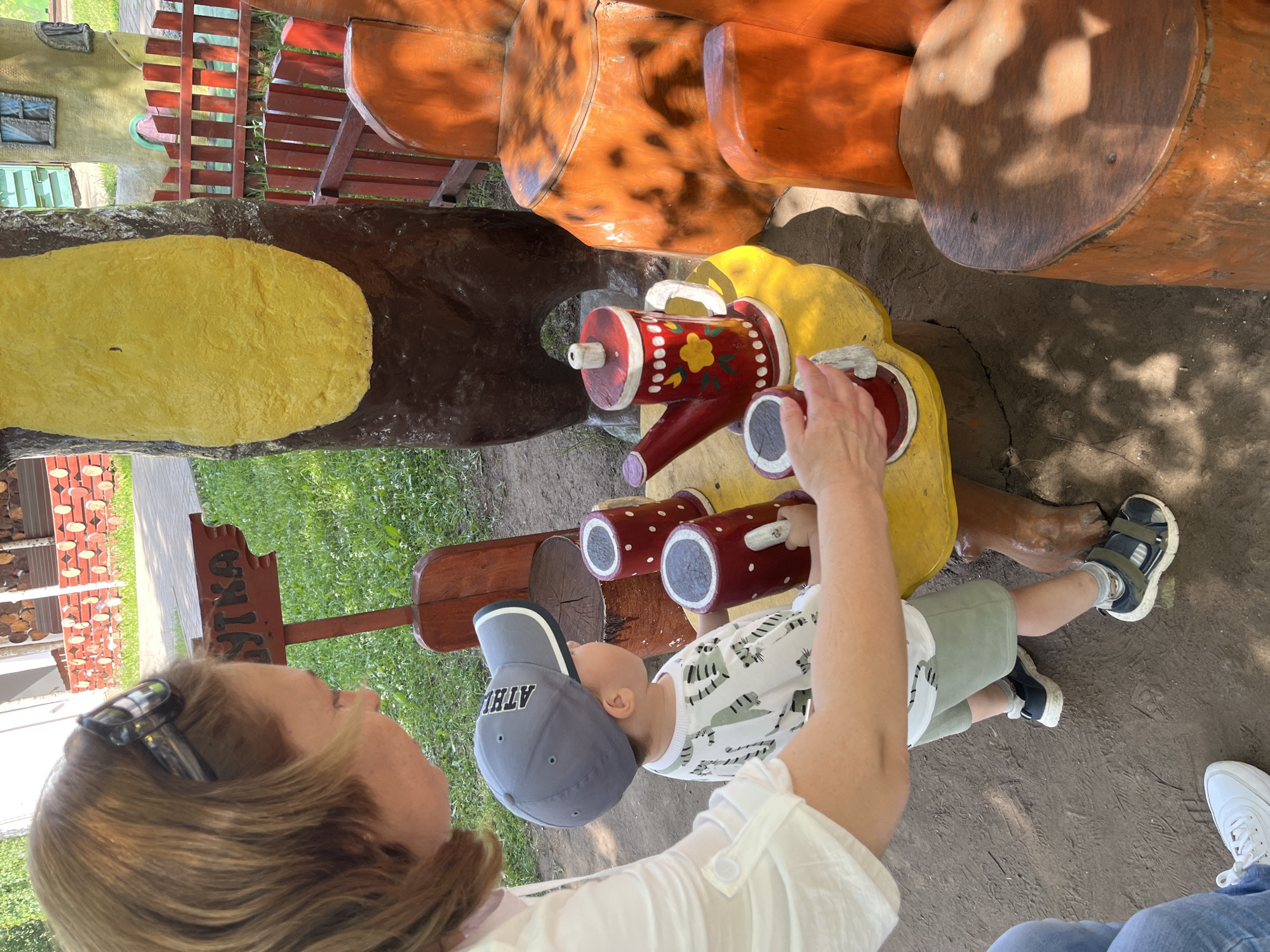 A woman with blonde hair and sunglasses is playing with a young child at a wooden table outside. The child, wearing a grey baseball cap, a white t-shirt with a green dinosaur print, and green shorts, is reaching for a red toy teapot on a yellow table. The table is supported by a brown, tree-like structure. There are several red toy cups on the table, and the woman's hand is reaching for one of them. In the background, a wooden fence and a building with a green door and windows are visible. The ground is dirt and scattered with grass and leaves. The image appears to be taken in an outdoor recreational area, possibly a children's play area, in the city of Chaikovsky, Russia, judging by the signage which includes Cyrillic lettering.