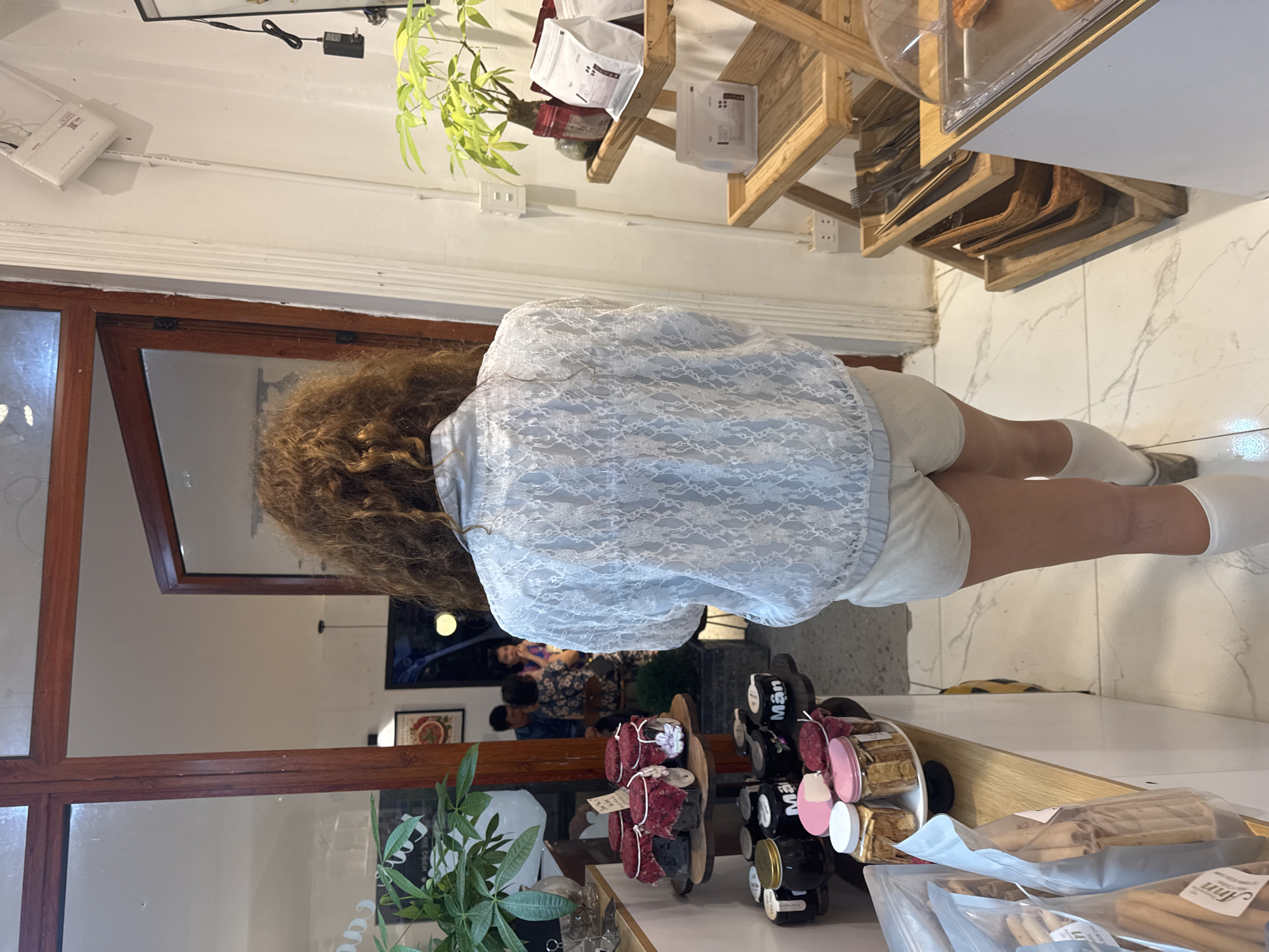 A woman with long, curly brown hair is standing with her back to the camera, facing a glass-paned door leading into a room. She is wearing a light blue lace jacket and light-colored shorts, with white socks pulled up to her knees. The background shows shelves filled with jars and bags of products. To the right, a wooden display shelf holds bags of coffee and other items. The floor is tiled with a marble pattern. The scene appears to be an indoor market or shop.