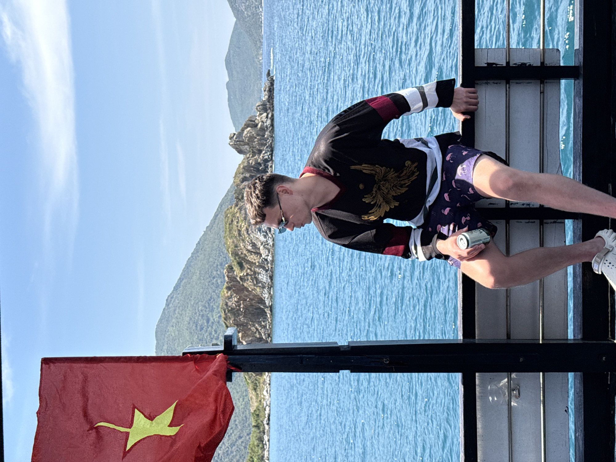 A man stands on a pier overlooking the sea and a rocky coastline. He wears a black long-sleeved shirt with a gold eagle design, flamingo-patterned shorts, and white Crocs. He holds a silver can in his right hand. A red flag with a gold star, likely the flag of Vietnam, is attached to a pier post on the left. The location is likely near Bac Nha Trang Ward, Vietnam, given the presence of the Vietnamese flag. The sky is blue with some clouds.