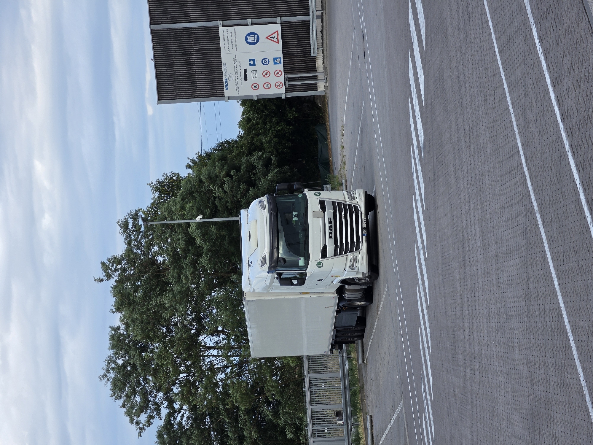 A white DAF truck is parked in a paved lot with white lane markings. The truck has a refrigerated trailer. To the left of the truck, there are green trees and a blue sky with white clouds. Behind the truck and to the right, there is a tall, dark-colored fence with a sign containing multiple pictograms and text. The text on the sign includes "ACHTUNG FUSSGÄNGER! LOOK OUT FOR PEDESTRIANS!" and "NAGEL-GROUP". Other visible text includes the truck's license plate "MOB 592" and the brand name "DAF" on the front of the truck. The scene appears to be outdoors during the daytime with fair weather. The parking lot suggests a commercial or industrial area, possibly a logistics hub or a truck stop. There are no people visible in the image.
