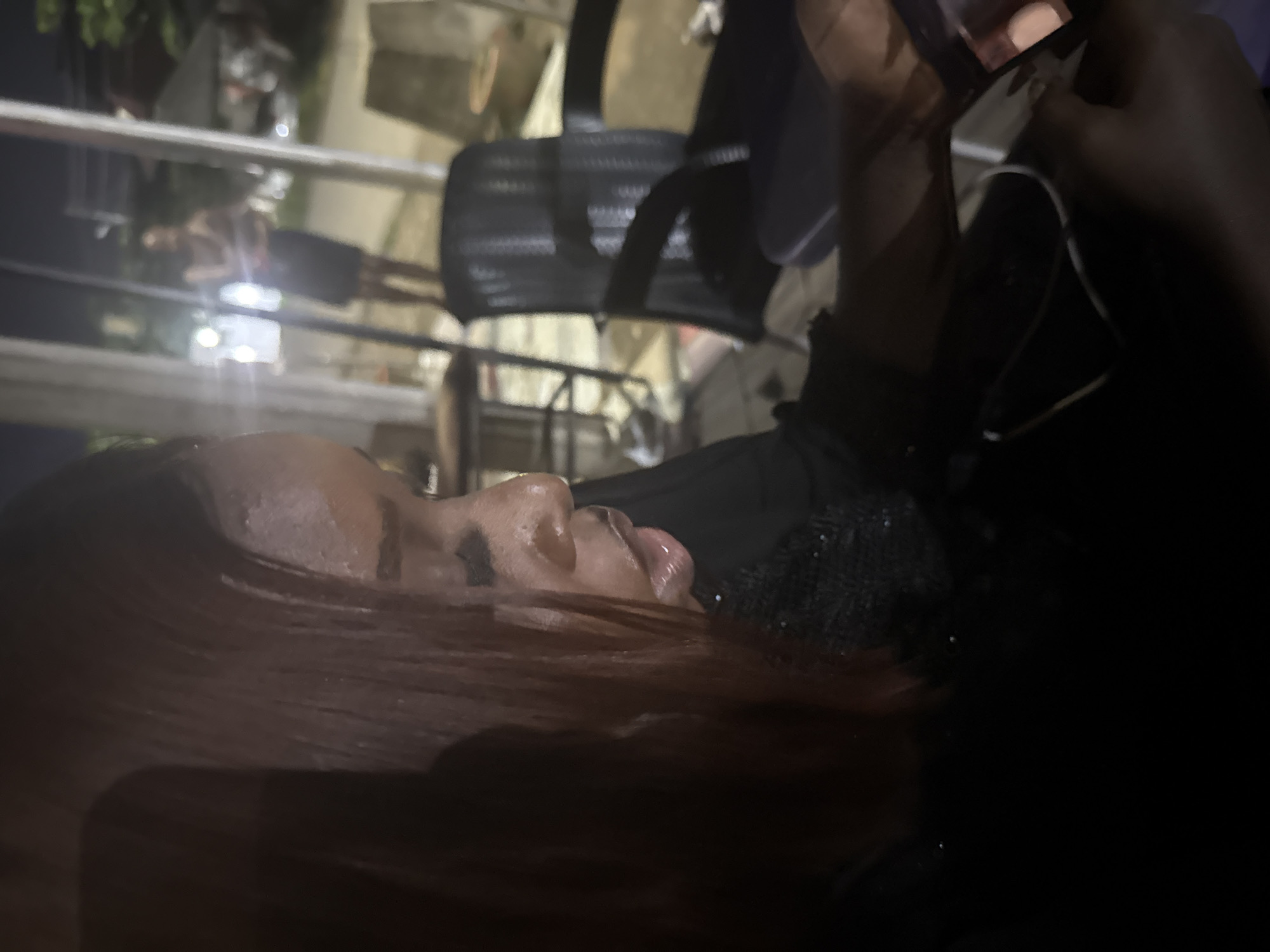 A close-up, side profile of a young Black woman's face is captured in low light, with her head turned to her right as she looks down at a mobile phone in her hands. Her auburn hair is visible, appearing slightly damp. She wears a black top with a textured neckline. In the background, out of focus, a person stands near a window, and to the right, a black office chair is visible. The setting appears to be an outdoor or semi-outdoor establishment at night, possibly a restaurant or bar, in Abuja, Nigeria.