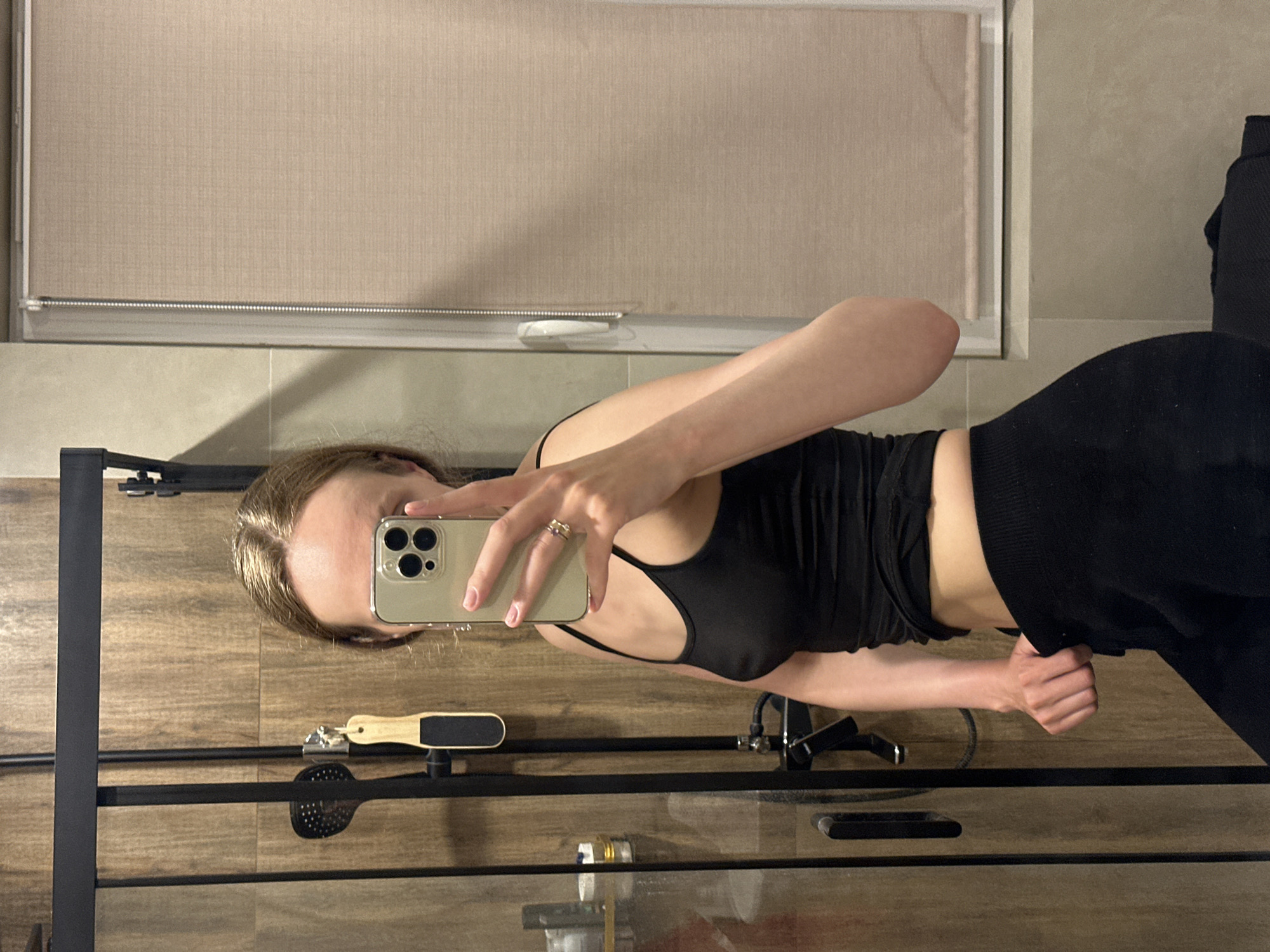 The image shows a person taking a selfie in a bathroom. The person is wearing a black tank top and black bottoms. The bathroom has wooden cabinets, a window with a beige blind, and a shower rack. A foot file and a shower head are visible on the rack. There is no indication of the location being in Подшивалово, Russia.