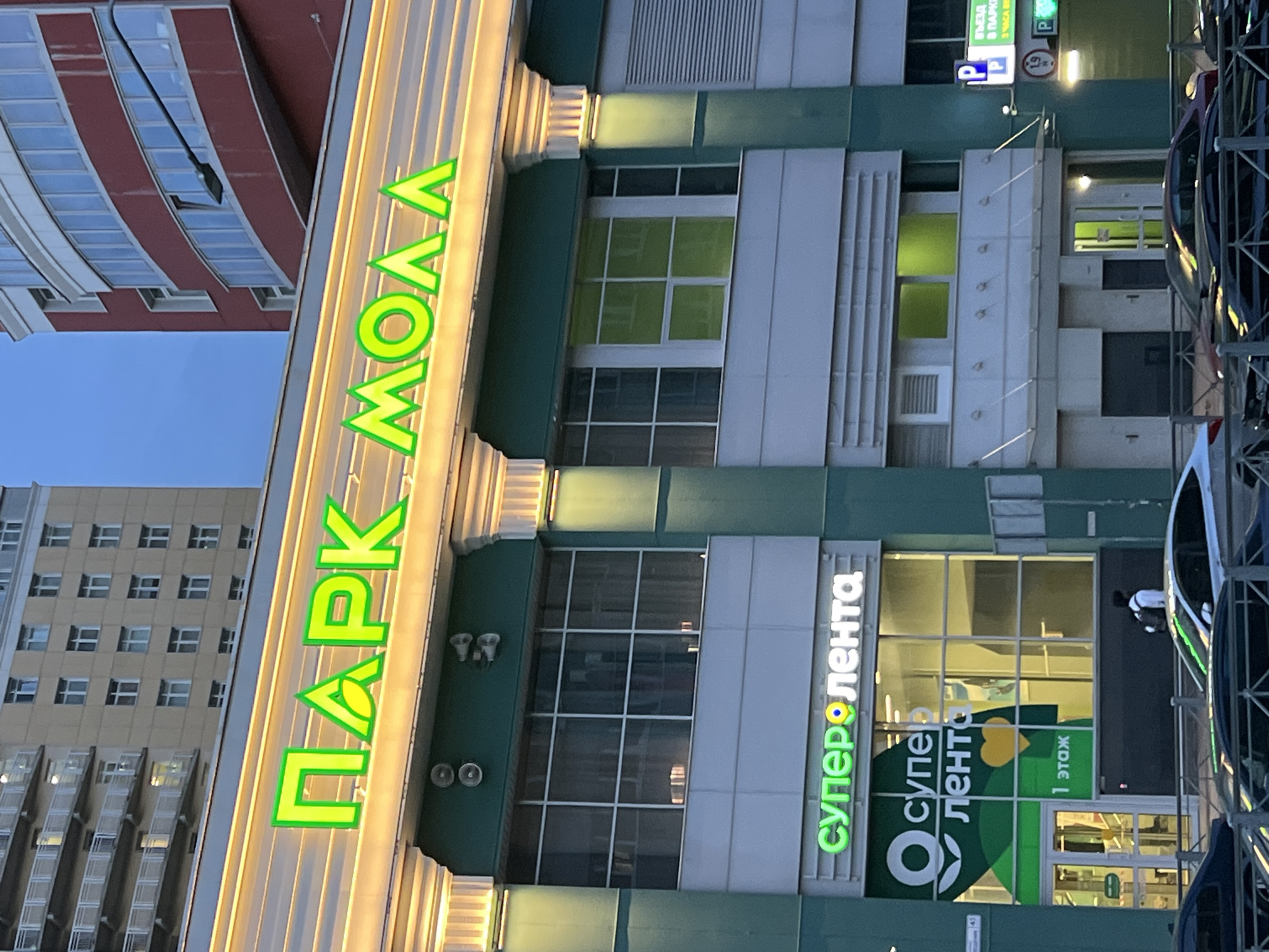 This media file captures the exterior of a modern building with a prominent sign in bright green neon letters that reads "ПАРК МОЛЛ" (PARK MALL). The building features a mix of architectural styles, with a red and white striped structure visible in the background, and a more contemporary design with large windows and metallic accents.
The "PARK MALL" sign is illuminated, suggesting it is either evening or an indoor setting with artificial lighting. Adjacent to the main sign, another green neon sign reads "Суперлента" (Superlenta), which appears to be a store or supermarket located within the mall. Below this, a logo with a green circle and heart shape is visible on a large window. There is also text in Russian indicating "1 этаж" (1st floor) and signs related to parking, with "P" symbols and Russian text that appears to translate to "ENTRY TO PARK" and "3 HOURS FREE".
One person is partially visible in the image, wearing a white shirt and dark pants, interacting with a window or display. The overall scene suggests a commercial area, possibly a shopping center or complex, with an emphasis on signage and branding. The lighting and colors create a vibrant and modern atmosphere.