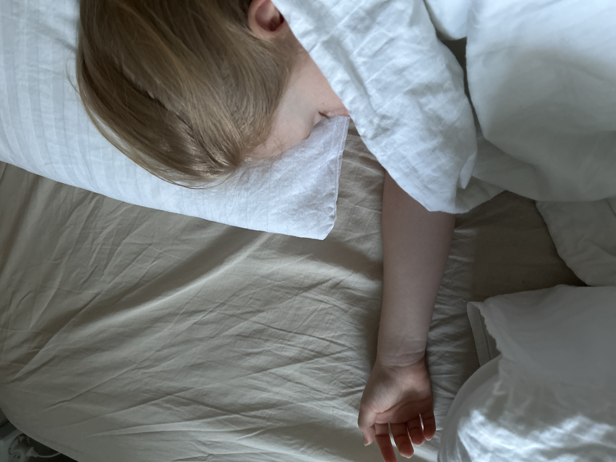 The image shows a person sleeping in a bed. The person has blonde hair and is lying on a white pillow, partially covered by a white blanket. One arm is extended out of the blanket and rests on the tan-colored bed sheet. There is a second white pillow near the edge of the bed. The scene appears to be indoors. The city of Chaikovsky, Russia, is not evident within the image.