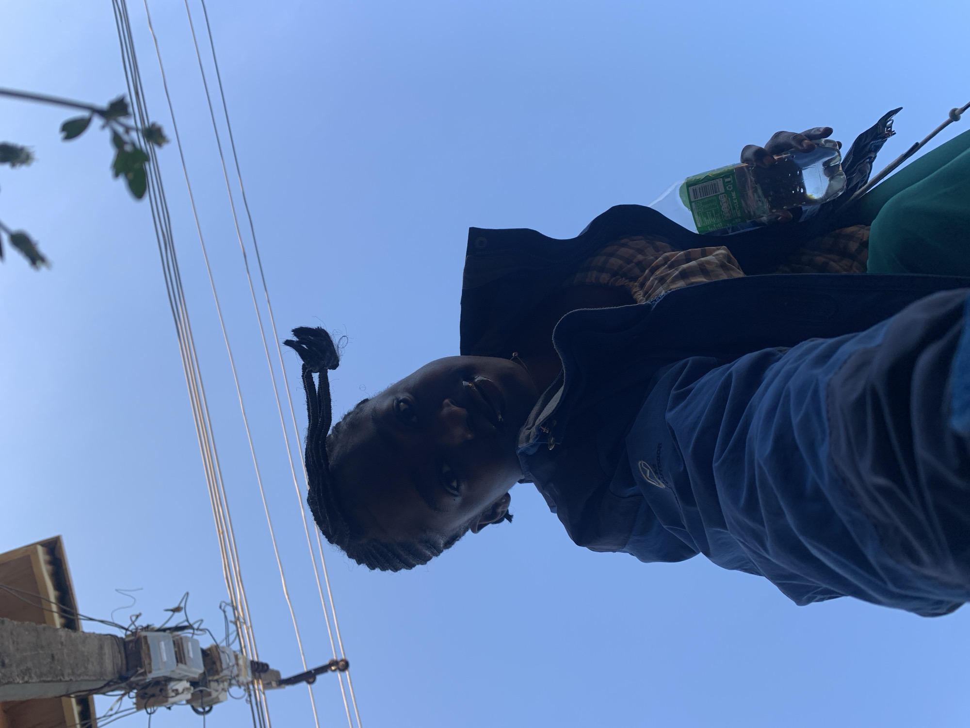 The image shows a young Black female, appearing to be in her late teens or early twenties, holding a plastic bottle of water. She is wearing a dark hooded jacket over a checkered top and green pants. Her hair is styled in braids, with one braid pulled back and secured with an elastic band. The perspective is from below, looking up at her against a clear blue sky, with power lines and a concrete pole with electrical equipment visible in the background. The setting is outdoors, likely in an urban or semi-urban environment given the presence of electrical infrastructure. There is no discernible information in the image that specifically places the location in Rafin Par, Nigeria.