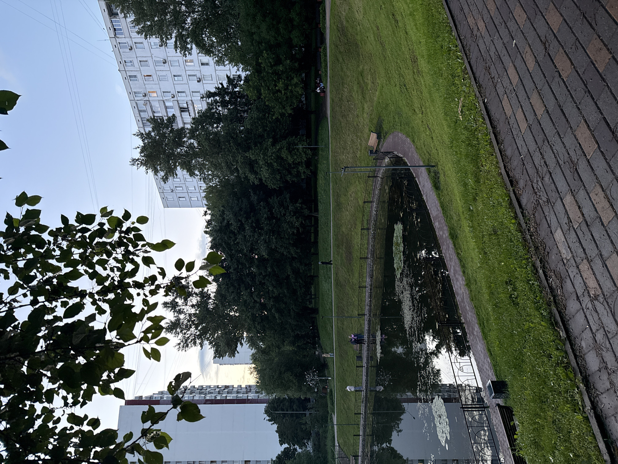 This image depicts a tranquil park scene in Moscow, Russia, featuring a small pond surrounded by lush greenery and residential buildings in the background. The pond, with lily pads scattered across its surface, reflects the overcast sky and the surrounding trees. A paved walkway with decorative fencing runs along the edge of the pond, where several individuals are casually strolling and conversing. Beyond the walkway, a gentle grassy slope leads up to a pathway lined with trees, where more people are seen enjoying the park. In the distance, tall, multi-story apartment buildings, characteristic of Moscow's urban landscape, dominate the skyline, some with lights on within their windows. The overall atmosphere is peaceful, suggesting a late afternoon or early evening in a typical residential area of the city.