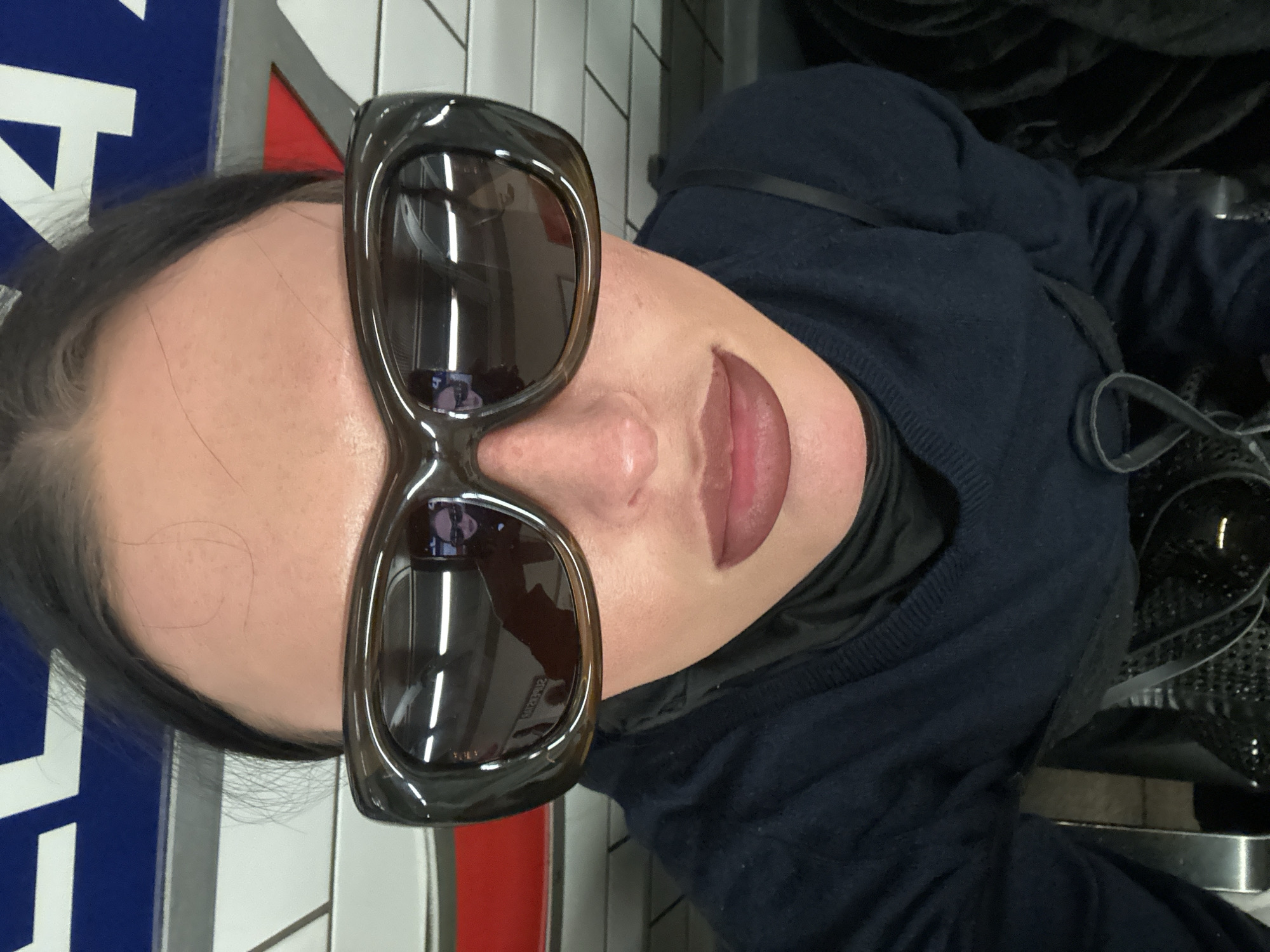A female individual is depicted in a close-up, upward-angled selfie, shown from the chest up. She has dark hair, prominent dark lip makeup, and wears oversized, dark brown, rectangular sunglasses. Her attire consists of a dark navy blue V-neck sweater layered over a darker garment. The sunglasses reflect the immediate environment, including elongated horizontal strip lights, a vague human figure, and structural elements consistent with a public transport setting. A black strap, likely from a bag, is visible towards the bottom right. The background features white rectangular ceramic tiles with dark grout lines. Above the tiled section, a blue horizontal sign band with white uppercase lettering is visible, showing partial characters resembling "L" on the left and "4" on the right. A red triangular shape is positioned below the "4". A horizontal red stripe runs along the wall beneath the blue sign. These distinctive architectural and signage elements indicate the location is within the London Underground system in the city of London, United Kingdom.