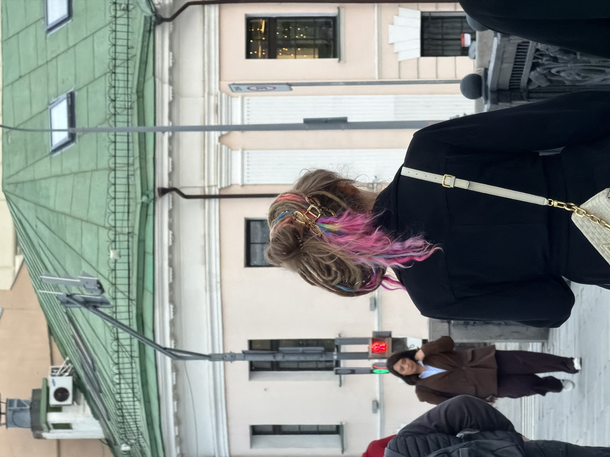 A street-level, eye-level medium shot depicts people walking on a sidewalk in an urban area, likely Moscow, Russia, suggested by architectural elements. A woman with dreadlocks that are partly pink and blue wears a black coat. She is walking to the right of the frame. Another woman, wearing a brown suit, is visible in the background. Traffic lights appear to be red for pedestrian crossing. Portions of building facades with varying architectural styles are also in view.