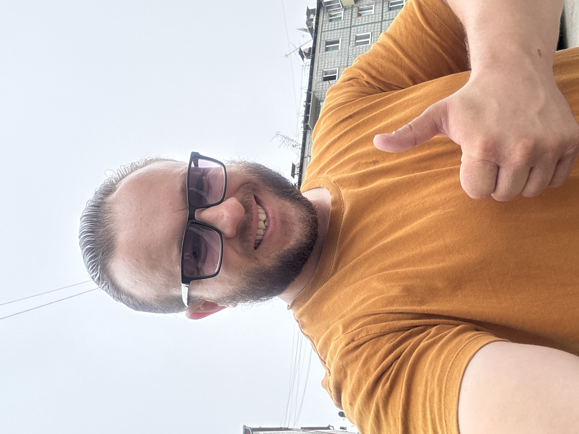 A selfie of a Caucasian man with light brown hair, a beard, and sunglasses is taken outdoors. He is wearing an orange t-shirt and giving a thumbs-up gesture. In the background, a building with a gray, checkered facade and multiple windows is visible, along with power lines and an overcast sky. The setting is consistent with an urban environment in Blagoveshchensk, Russia, given the architecture of the building and the presence of overhead electrical wires.