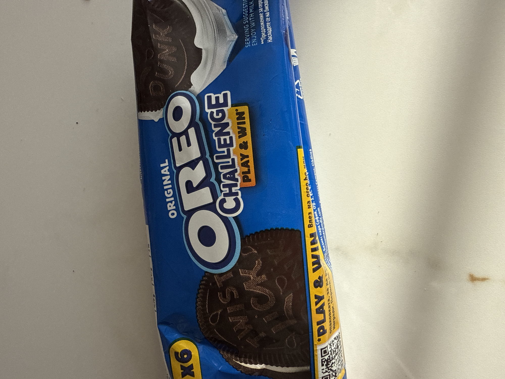 An overhead photograph depicts a blue package of "ORIGINAL OREO CHALLENGE" cookies resting on a light-colored, off-white horizontal surface. The package, oriented horizontally, displays "X6" in a yellow oval on its top-left corner, indicating the quantity. The main branding "OREO" is prominently centered in white block letters with a light blue outline. Below "CHALLENGE," a yellow box contains "PLAY & WIN."
To the left, a chocolate cookie with a white cream filling is partially visible, embossed with the words "TWIST LICK." To the right, another chocolate cookie, also partially visible, is embossed with "DUNK" and appears to be dipped into a depiction of white milk. Small text in white states "SERVING SUGGESTION" and "ENJOY WITH MILK," followed by a Cyrillic translation: "Предложение за сервиране. Насладете се на бисквитките с мляко."
Along the bottom of the package, a yellow and blue strip features a QR code on the left and the repeated "PLAY & WIN." Partially visible Cyrillic text translates to details about the game, including "Влез на играта..." (Enter the game...) and instructions to register a code from the package to win prizes. Smaller English text identifies "Business Park Sofia, str. 3" and "consumer@mdlz.com," with additional product information partially obscured.
The surface below the package has a faint, irregular brown mark in the lower central area. The background above the package is a plain, light-colored wall or vertical surface with a few minute dark specks. No individuals or active interactions are present in the image. The location is an indoor setting in Unknown, Unknown.