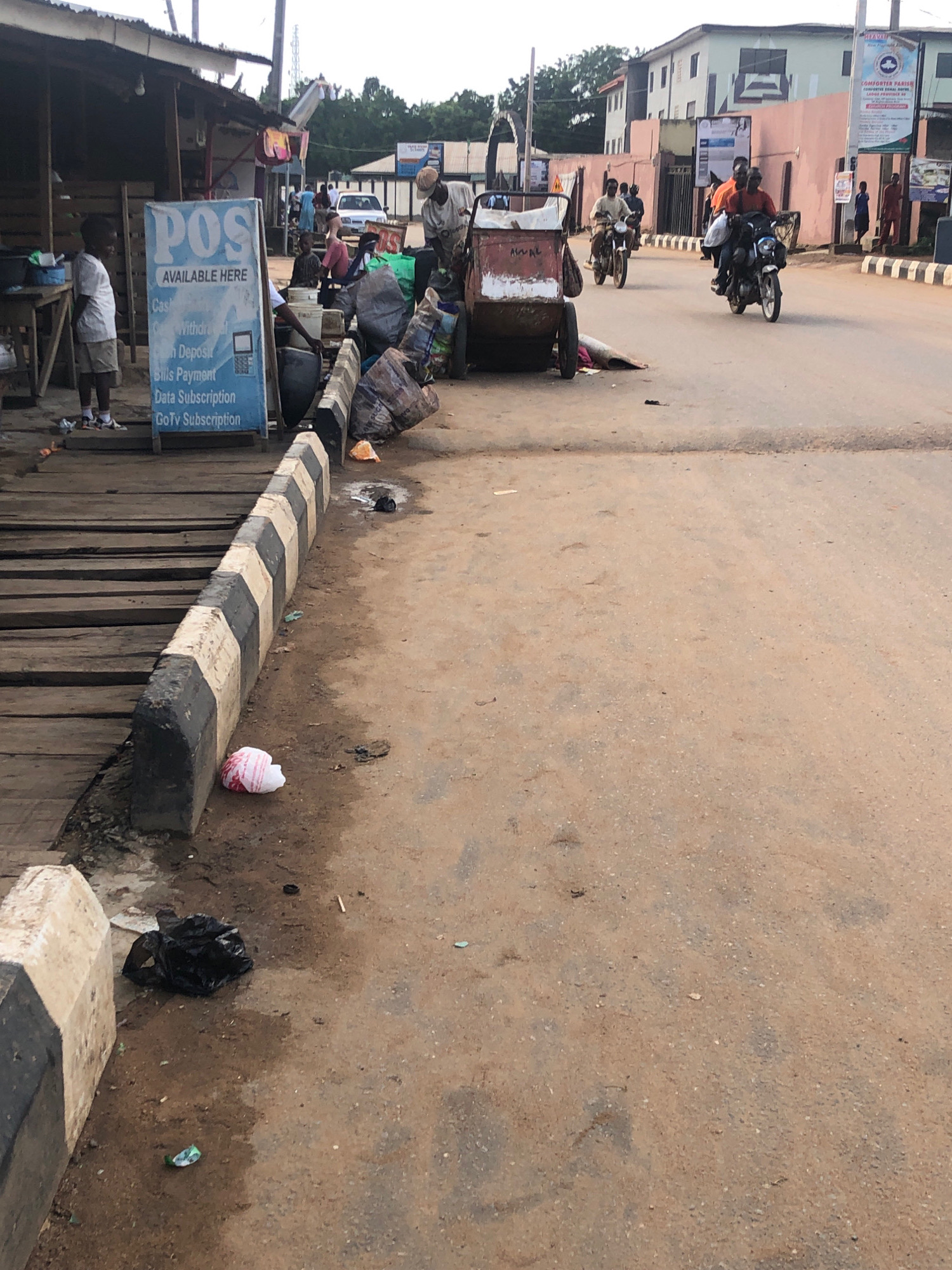 The image depicts a street scene in Lagos, Nigeria. In the foreground, a wooden walkway with black and white striped borders lines the street. On the walkway, a young boy in a white shirt and shorts stands to the left, facing away from the camera. A blue sign advertising "POS AVAILABLE HERE" with services like cash withdrawal, deposit, and subscriptions is prominent. Further down the walkway, a man is interacting with a cart loaded with various items, including bags and what appears to be a large white bucket.
The street itself is paved with asphalt and shows signs of wear. Several motorcycles are present. One motorcycle carrying two people in orange shirts is in the mid-ground, heading towards the right. Another motorcycle with a rider in a light-colored shirt is seen further back. A parked cart, possibly a wheelbarrow, is also visible on the left side of the street.
The background features buildings, including a pink building with signs and a white building. The sky is overcast. Litter is visible on the street and the edge of the walkway, including a black plastic bag and a crumpled white item near the border. The overall impression is of a busy, somewhat informal commercial area.
