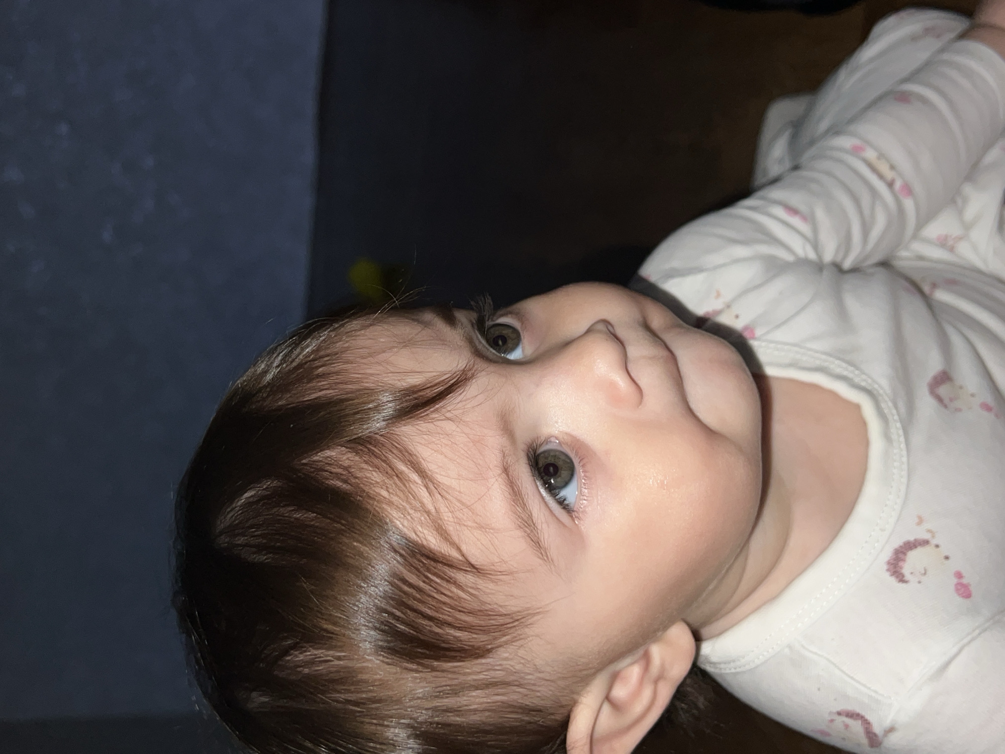 A close-up portrait in Elena, Bulgaria, shows an infant or toddler with brown hair and hazel eyes looking towards the upper right. The child's lips are slightly pursed, and the left ear is visible. The subject wears a white, long-sleeved garment patterned with light pink or brown animal designs. The background is dark and out of focus, featuring a muted blue-grey upper section and a darker brown lower section, with a faint yellow blur present in the upper right.