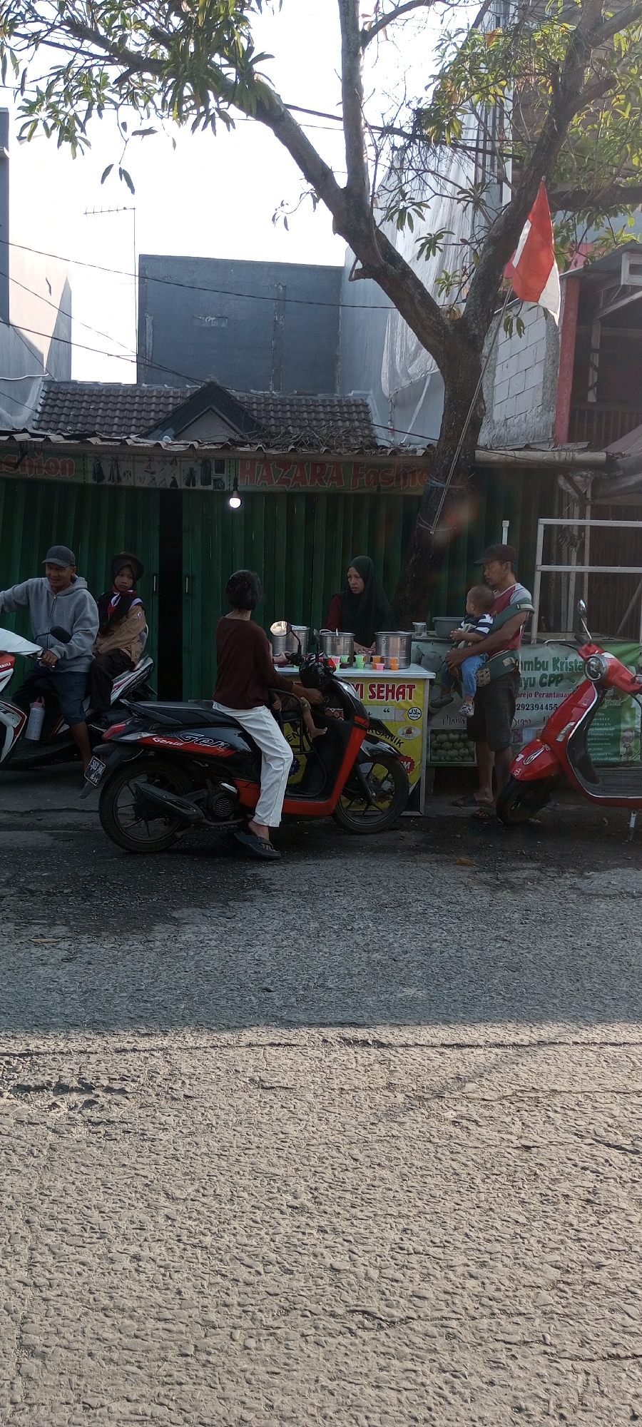 In Setu, Indonesia, on a paved street with a textured surface, a scene unfolds in front of a green storefront with the signage "HAZARA Fashion". The time of day appears to be late afternoon, indicated by the long shadows cast by the trees and buildings.
Several people are present. On the left, a man wearing a grey hoodie and a black baseball cap is seated on a white motorcycle, looking forward. Beside him on the same motorcycle, a young person in a brown outfit and a matching head covering is seated.
In the center, a person wearing a dark brown top and white pants is seated on a red and black motorcycle, facing a food stall. This stall features a white counter with several metal containers and small cups of colored beverages. A woman wearing a black hijab and a red top stands behind the counter, serving customers.
To the right of the food stall, a man holding a baby is standing. The baby is wearing a blue and white striped outfit. A red scooter is parked next to them. The background includes a tree with a red and white flag attached to one of its branches, and a building with a tiled roof. The overall atmosphere suggests a casual, everyday scene with people engaging in commerce and daily activities.